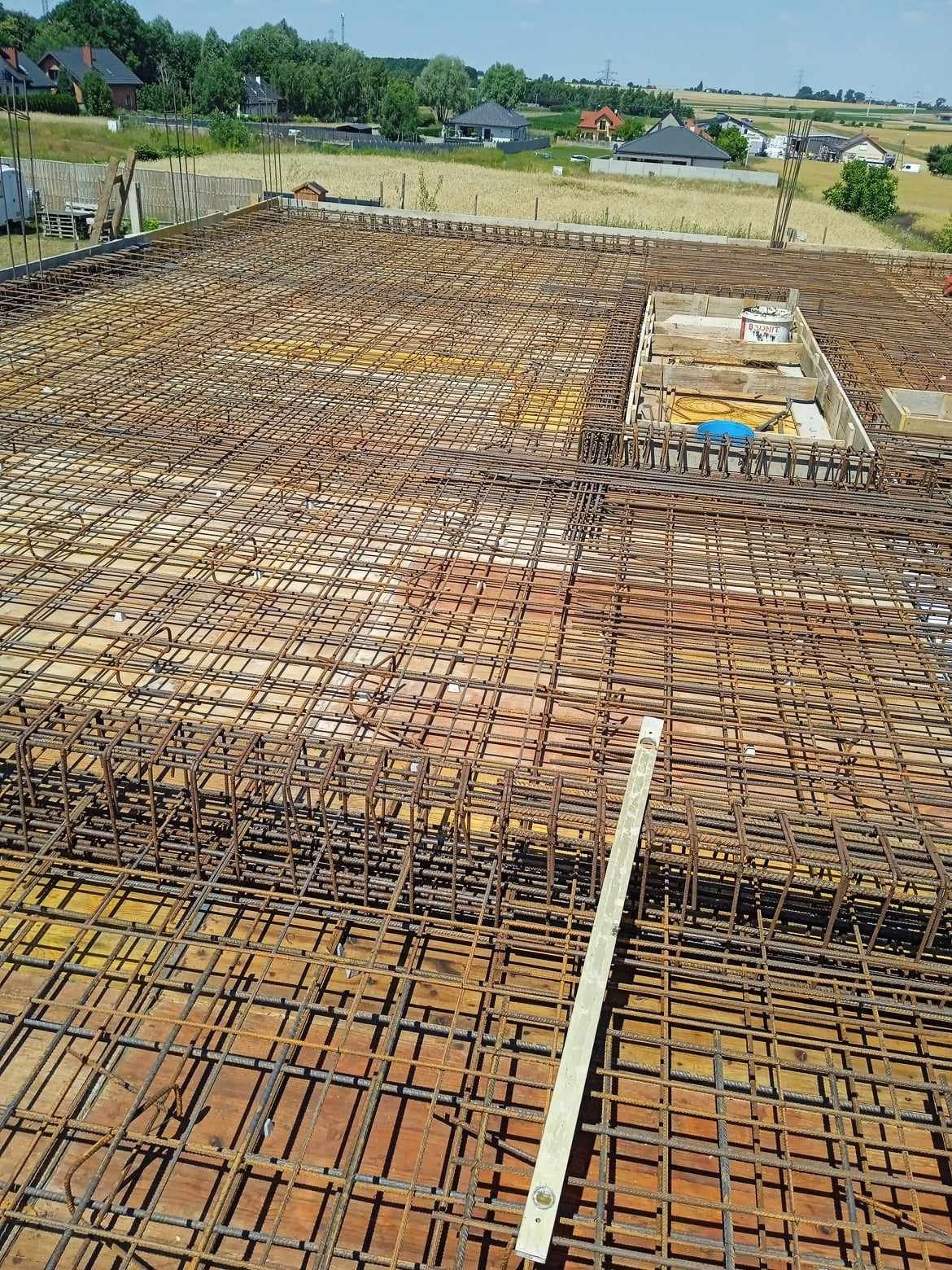 Zbrojenie płyty fundamentowej z widocznym szalunkiem na otwór instalacyjny i poziomicą, w tle zabudowa jednorodzinna.