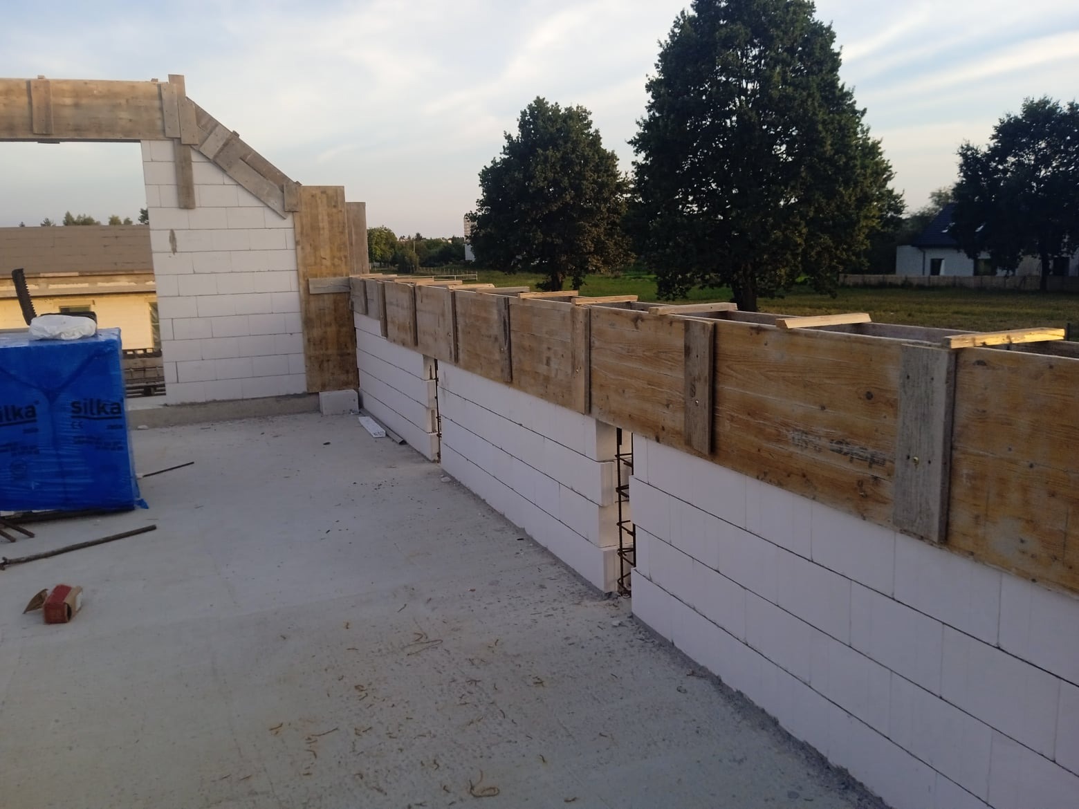 Budowa domu: Ściany z bloczków silikatowych z drewnianym szalunkiem, widoczne zbrojenia i paleta z materiałami budowlanymi w tle.