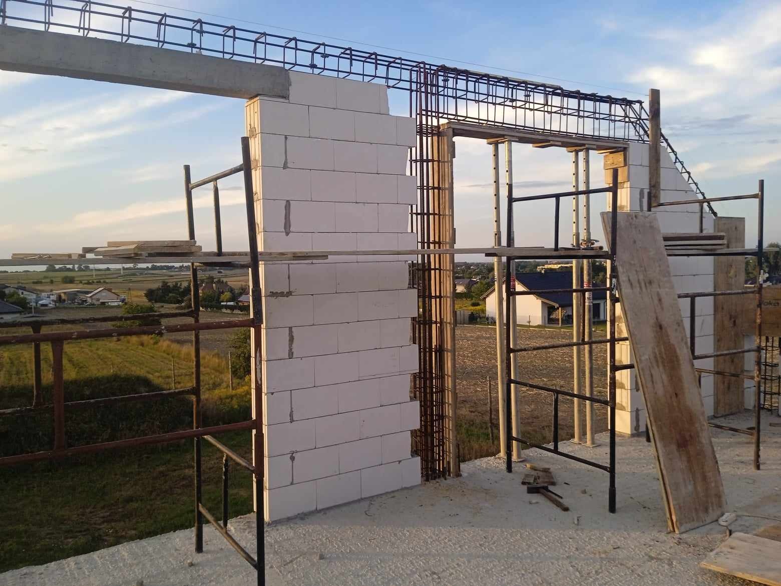 Wznoszenie ścian z białych bloczków na budowie domu jednorodzinnego, widoczne zbrojenie i rusztowania, w tle krajobraz wiejski.