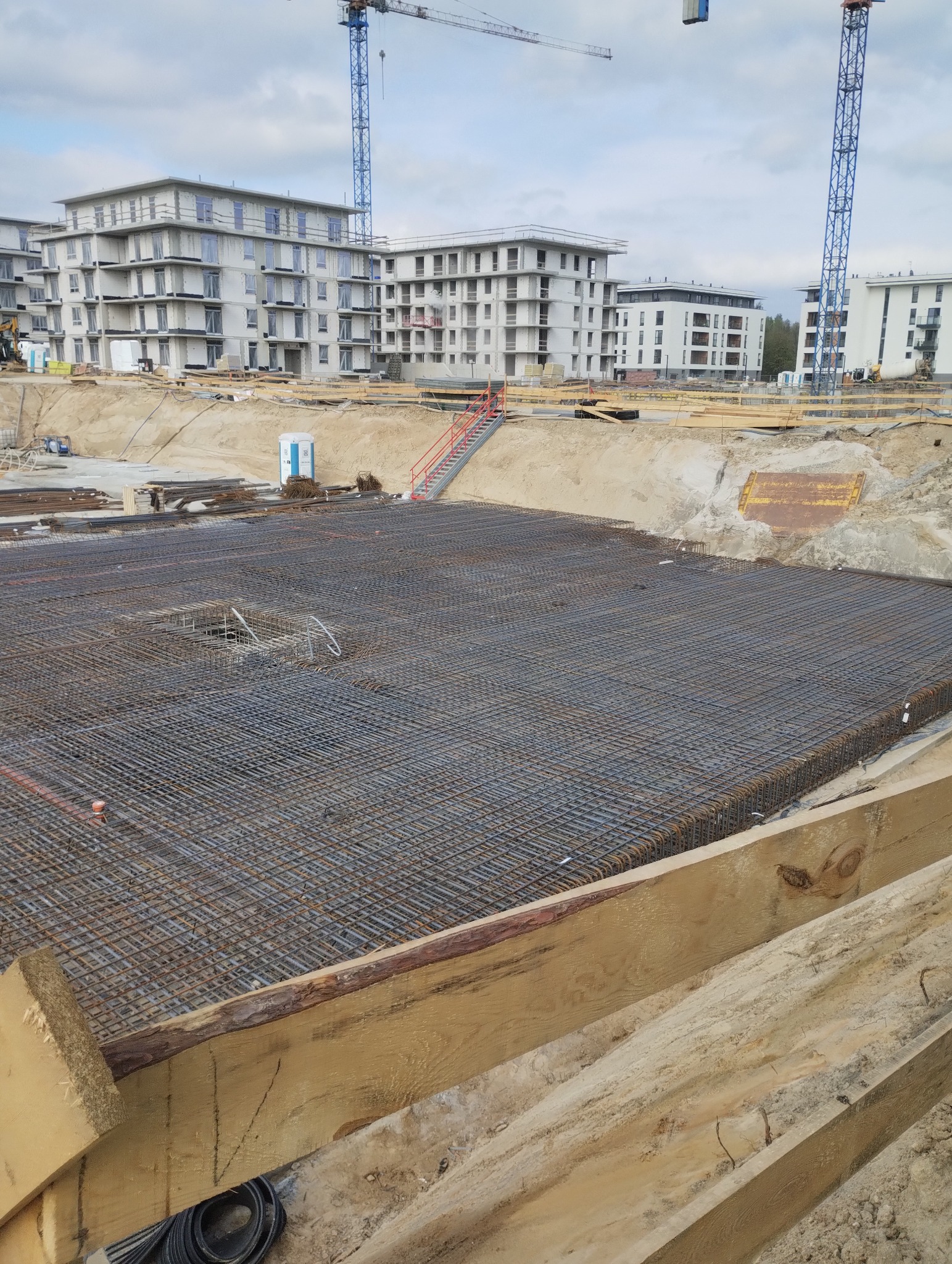 Siatka zbrojeniowa ułożona na budowie pod przyszły fundament, w tle surowe budynki mieszkalne i dźwigi