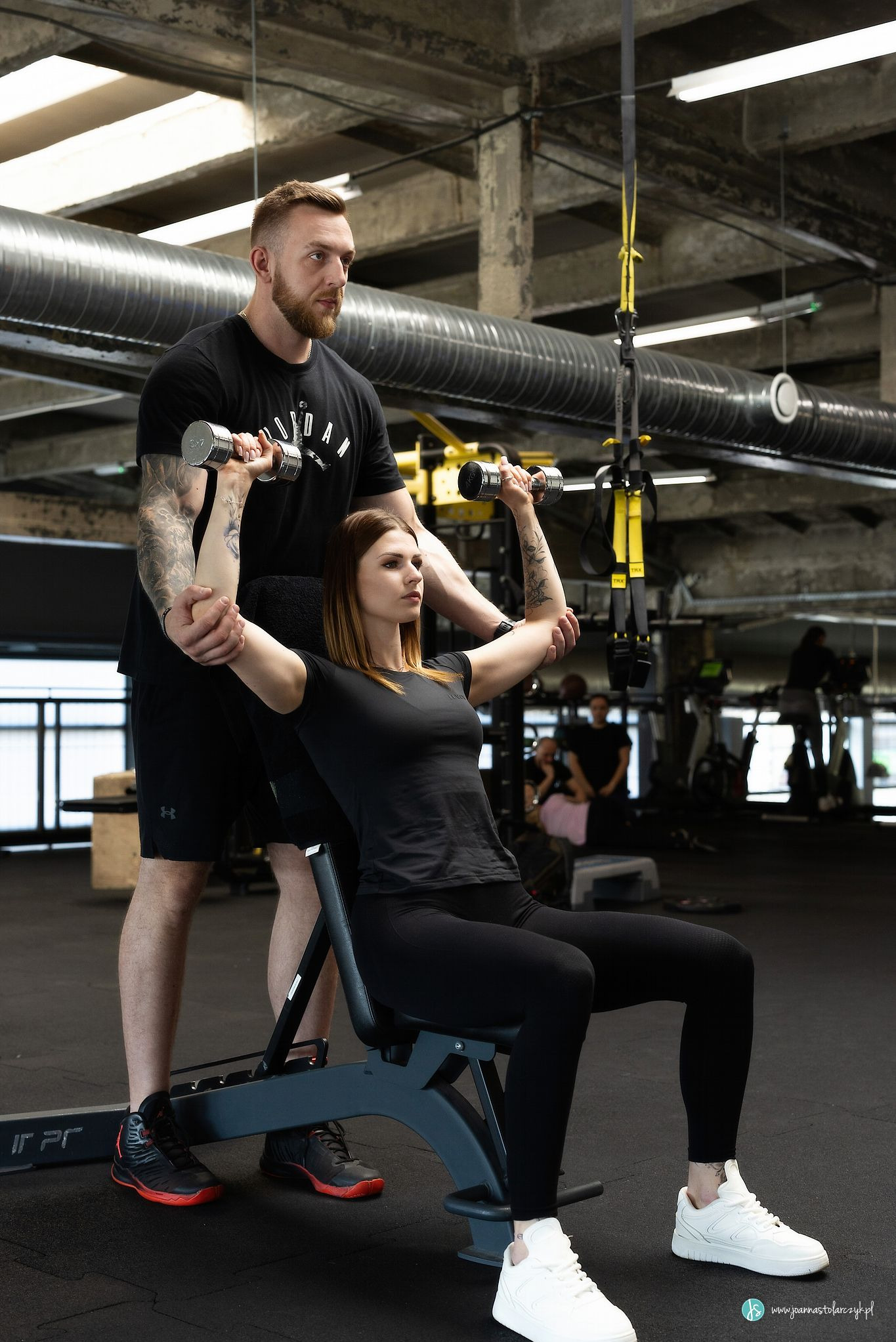 Kobieta ćwiczy unoszenie hantli siedząc na ławce pod nadzorem mężczyzny z tatuażami w siłowni o surowym wystroju.