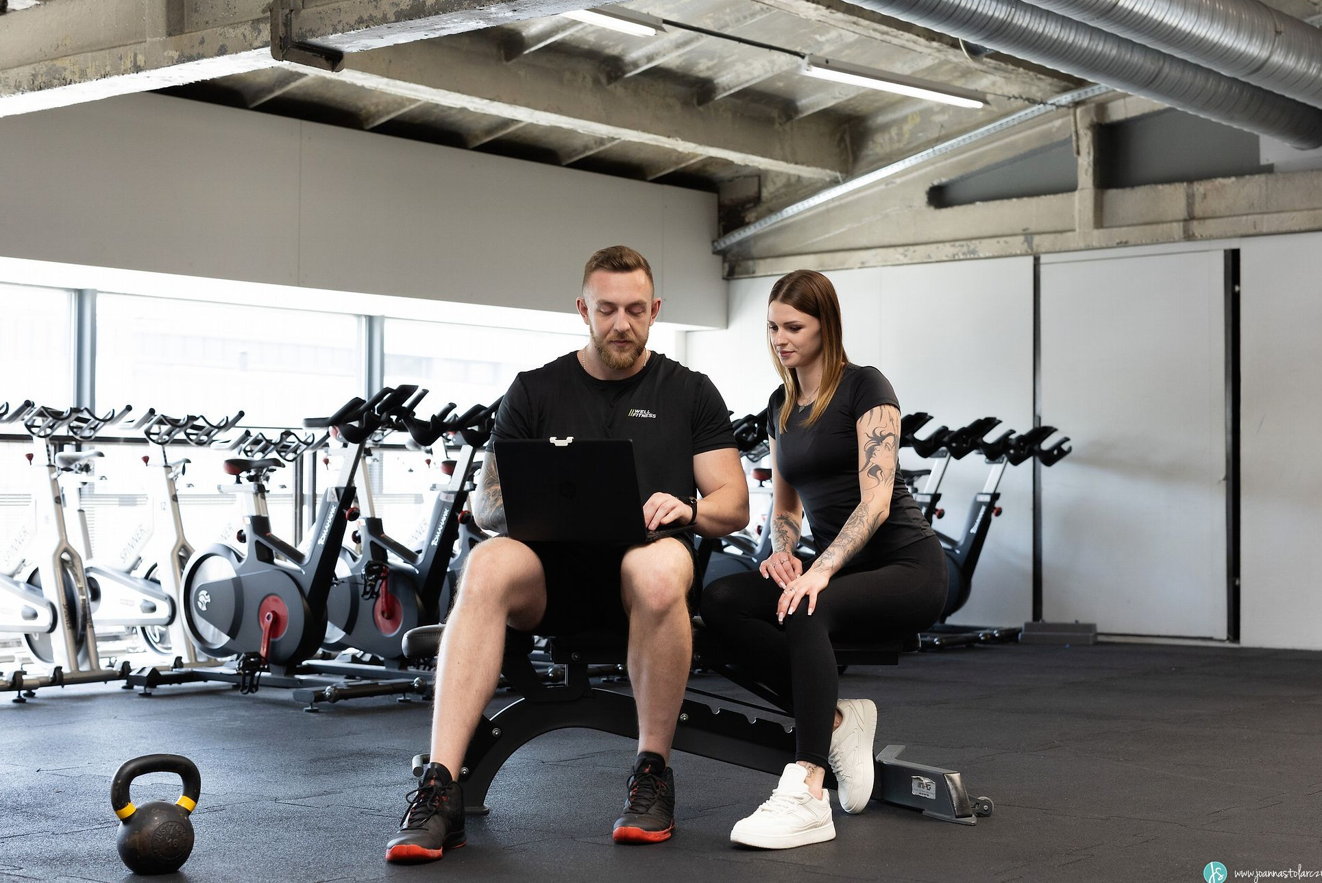 Mężczyzna z brodą w czarnej koszulce z logo Well Fitness siedzi na ławeczce w siłowni, korzystając z laptopa, obok siedzi kobieta z tatuażami, w tle rowery stacjonarne i odważnik kettlebell.