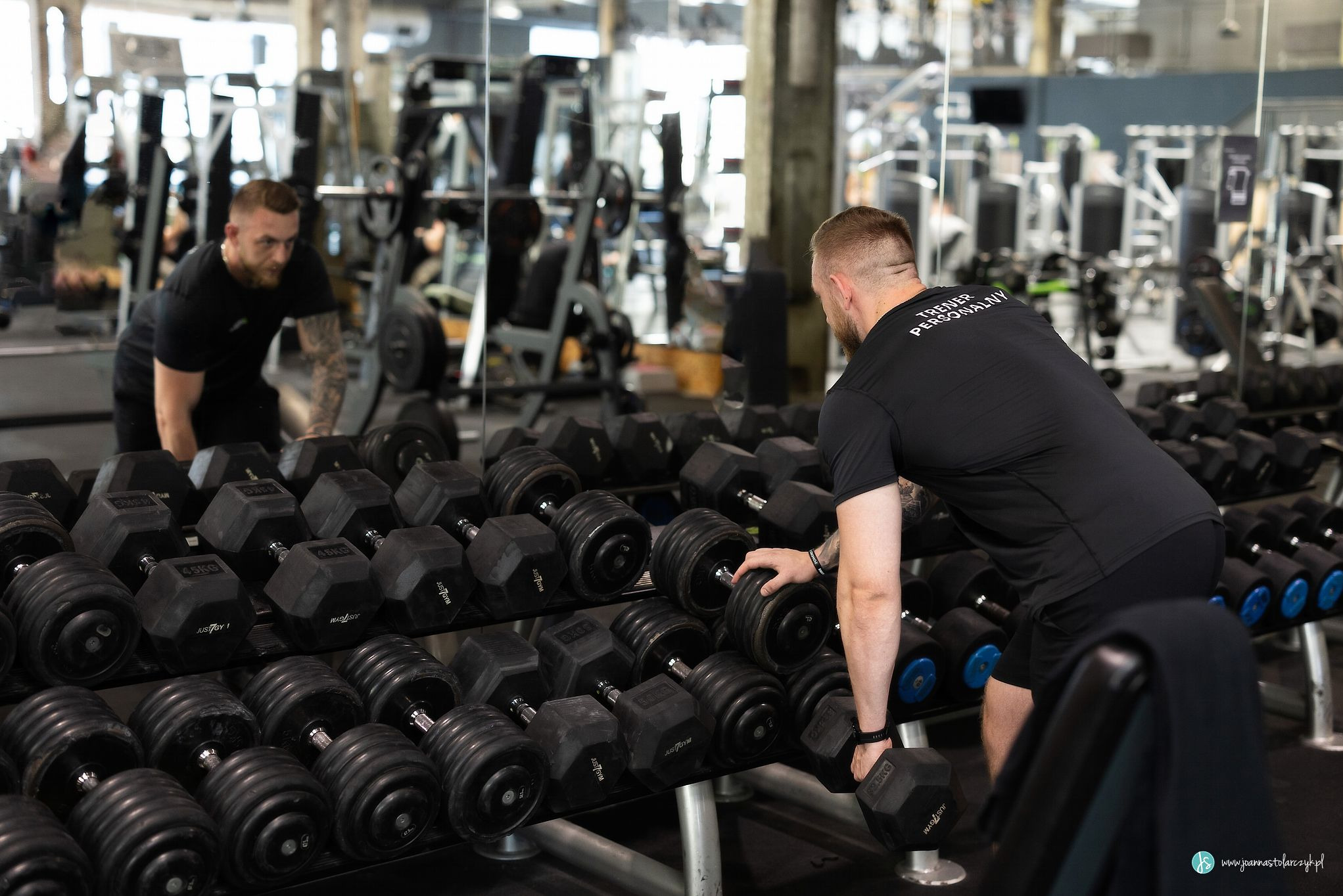 Mężczyzna w czarnej koszulce z napisem 'Trener Personalny' na plecach sięga po hantle w siłowni, widoczne rzędy ciężarków i lustrzane odbicie.