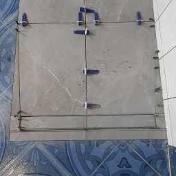 Masters Gres Jan Zonenberg - Układanie szarych płytek podłogowych z użyciem niebieskich klipsów poziomujących i metalowych listew, widoczne nogi w zabrudzonych butach roboczych oraz fragment niebieskiej, wzorzystej podłogi.