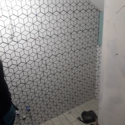 Masters Gres Jan Zonenberg - Ściana łazienki w trakcie układania białych płytek o wzorze kostki 3D z ciemnymi fugami, widoczne narzędzia i fragment instalacji hydraulicznej, podłoga wyłożona podłużnymi płytkami imitującymi...