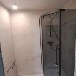 Nowoczesna kabina prysznicowa z czarnymi profilami w jasnej łazience po remoncie, widoczne płytki i narzędzia, taśma malarska na podłodze.