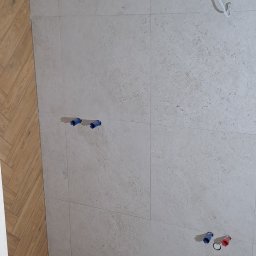 Wykończona strefa prysznicowa z odpływem liniowym i drewnopodobnymi płytkami ułożonymi w jodełkę, kontrastującymi z jasnymi, kwadratowymi płytkami na ścianach.