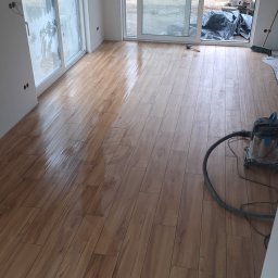 Masters Gres Jan Zonenberg - Jasne pomieszczenie z nowo ułożoną, błyszczącą podłogą z płytek imitujących drewno. Widoczne duże drzwi tarasowe i plac budowy z koparką na zewnątrz. Wnętrze w trakcie prac wykończeniowych.