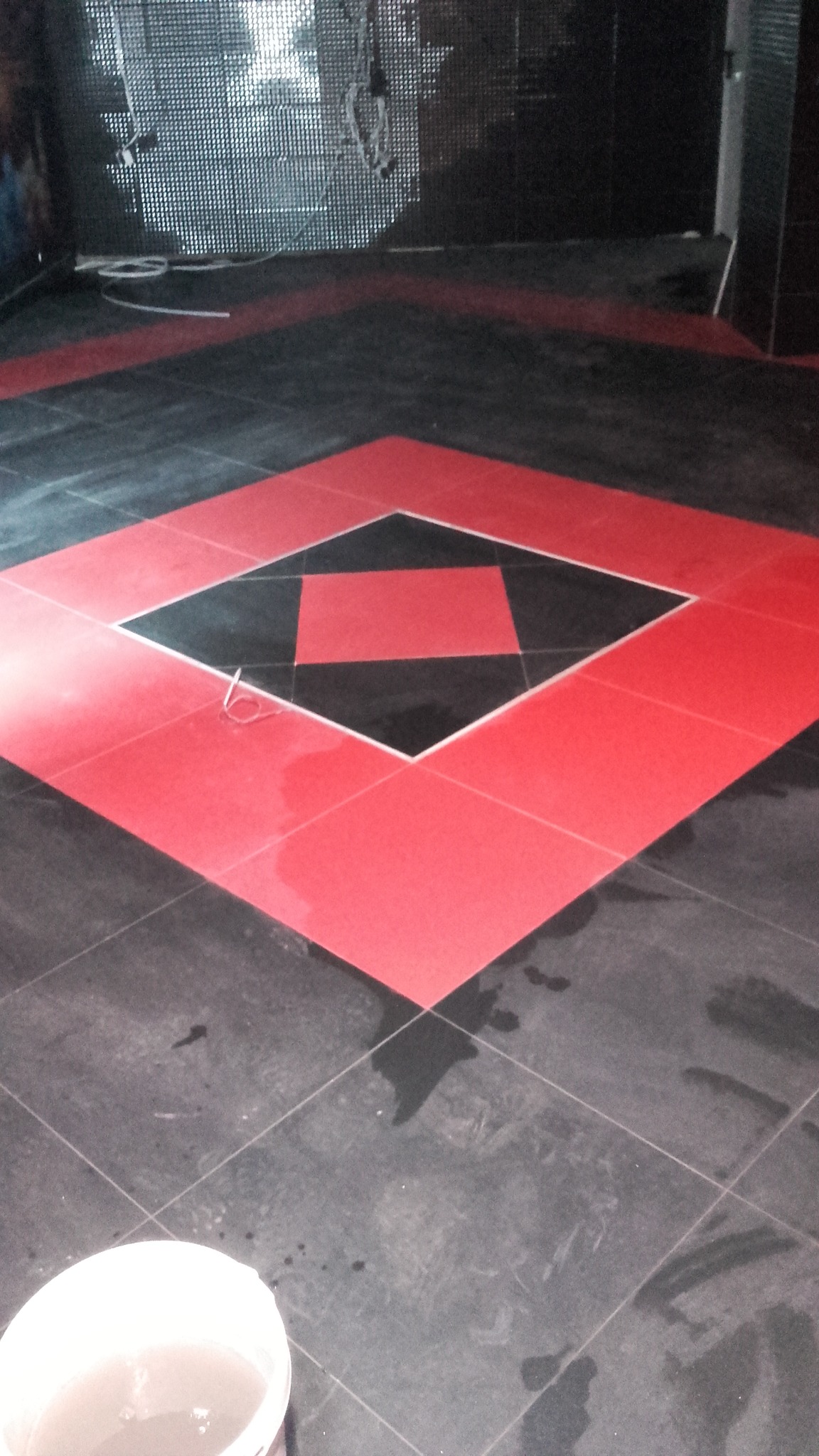 Ułożona pod kątem posadzka z czarno-czerwonych płytek, widoczny fragment białego wiadra z wodą w rogu, ślady po pracy na płytkach.