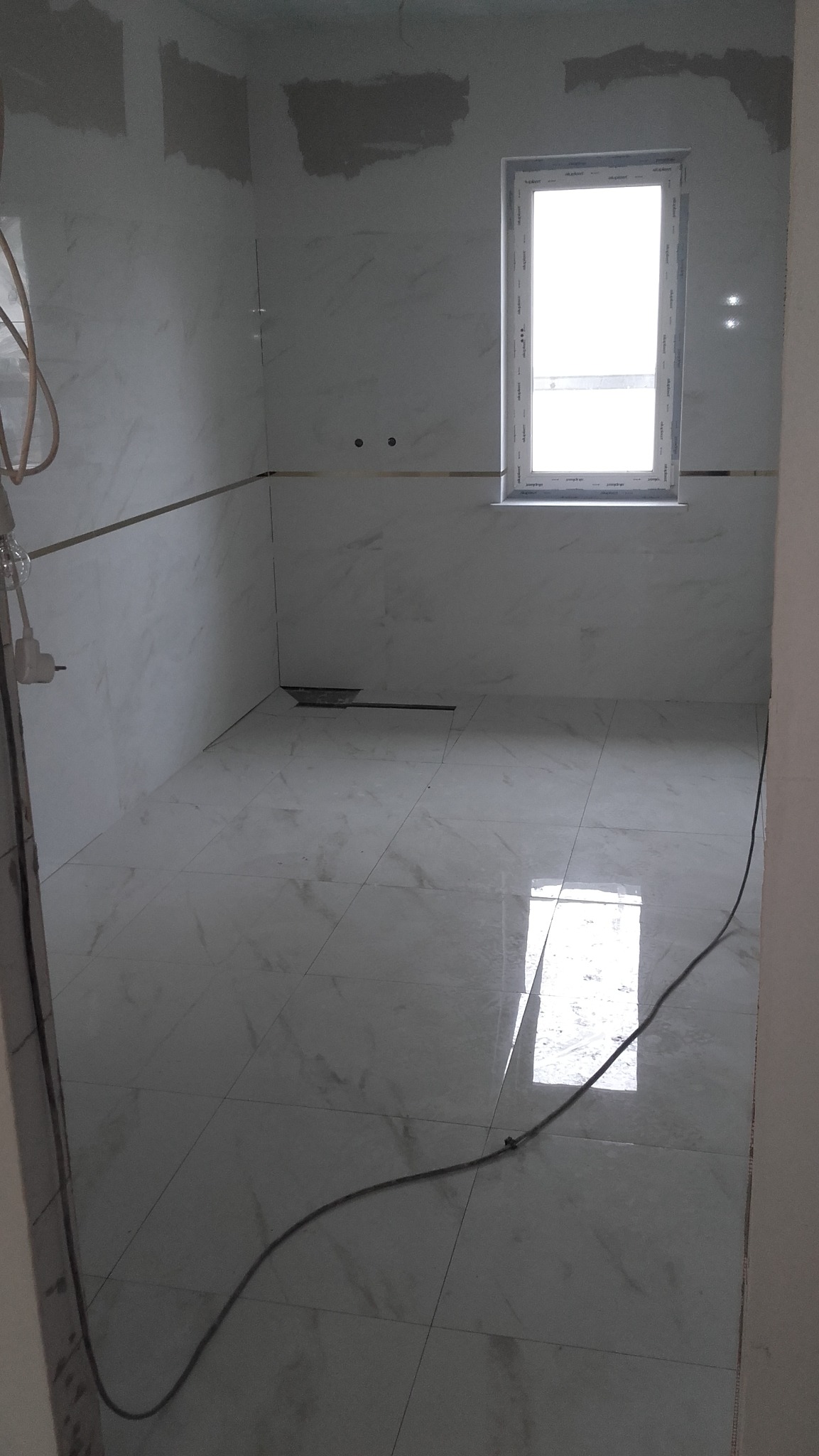 Wykończona łazienka z białymi marmurkowymi płytkami na ścianach i podłodze, z widocznym odpływem liniowym i oknem.