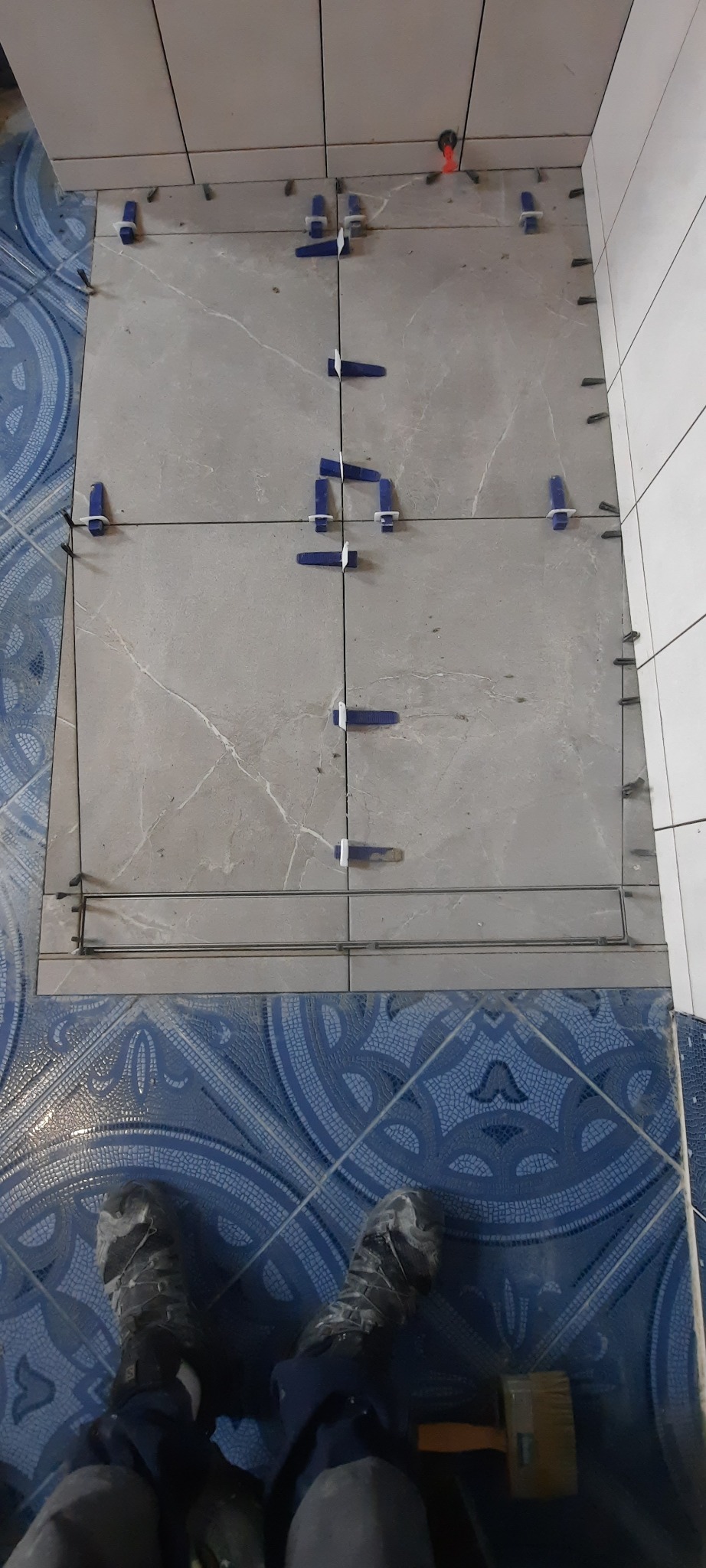 Układanie szarych płytek podłogowych z użyciem niebieskich klipsów poziomujących i metalowych listew, widoczne nogi w zabrudzonych butach roboczych oraz fragment niebieskiej, wzorzystej podłogi.