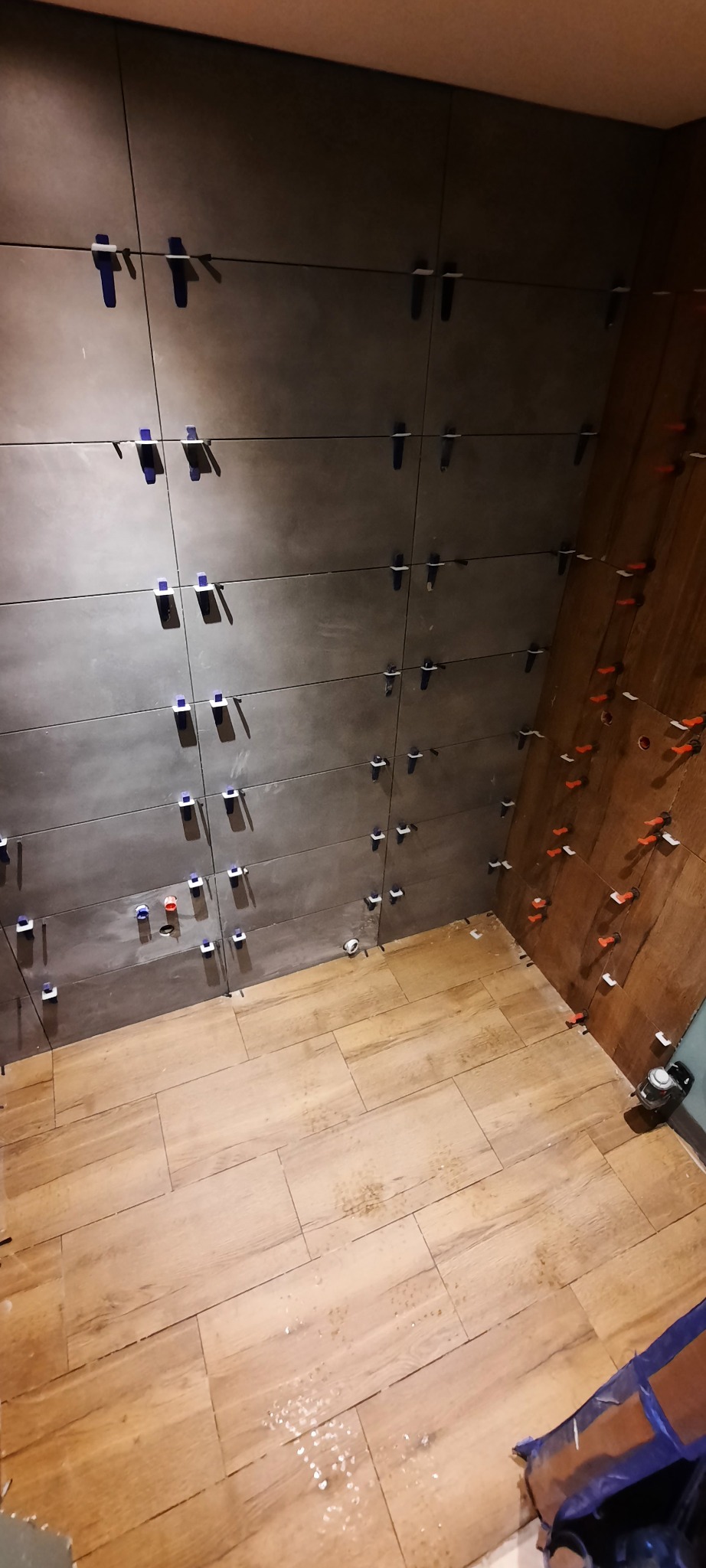 Montaż ciemnoszarych płytek ściennych z użyciem niebieskich i czerwonych krzyżyków dystansowych, widoczne płytki podłogowe imitujące drewno, laserowy niwelator przy ścianie.