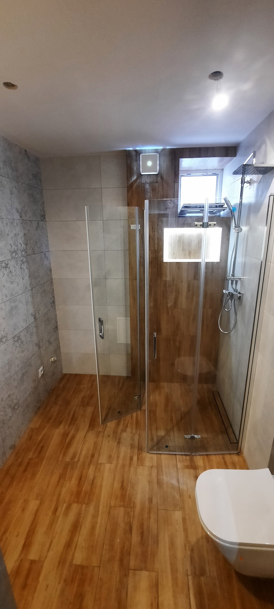 Nowoczesna łazienka z kabiną prysznicową typu walk-in, drewnopodobną podłogą, szarą płytką imitującą kamień na ścianie oraz podwieszaną toaletą.