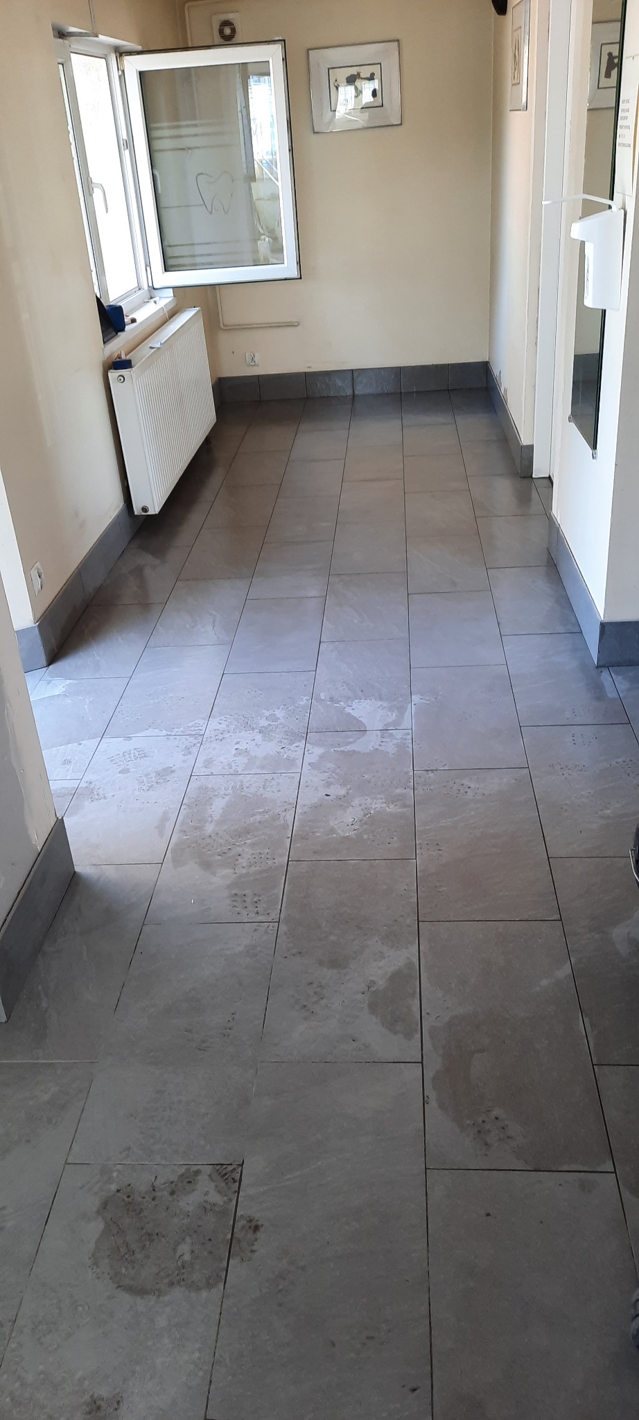 Świeżo ułożona, szara podłoga z płytek w pomieszczeniu, prawdopodobnie w gabinecie dentystycznym, z widocznymi śladami po czyszczeniu i oknem z rysunkiem zęba.