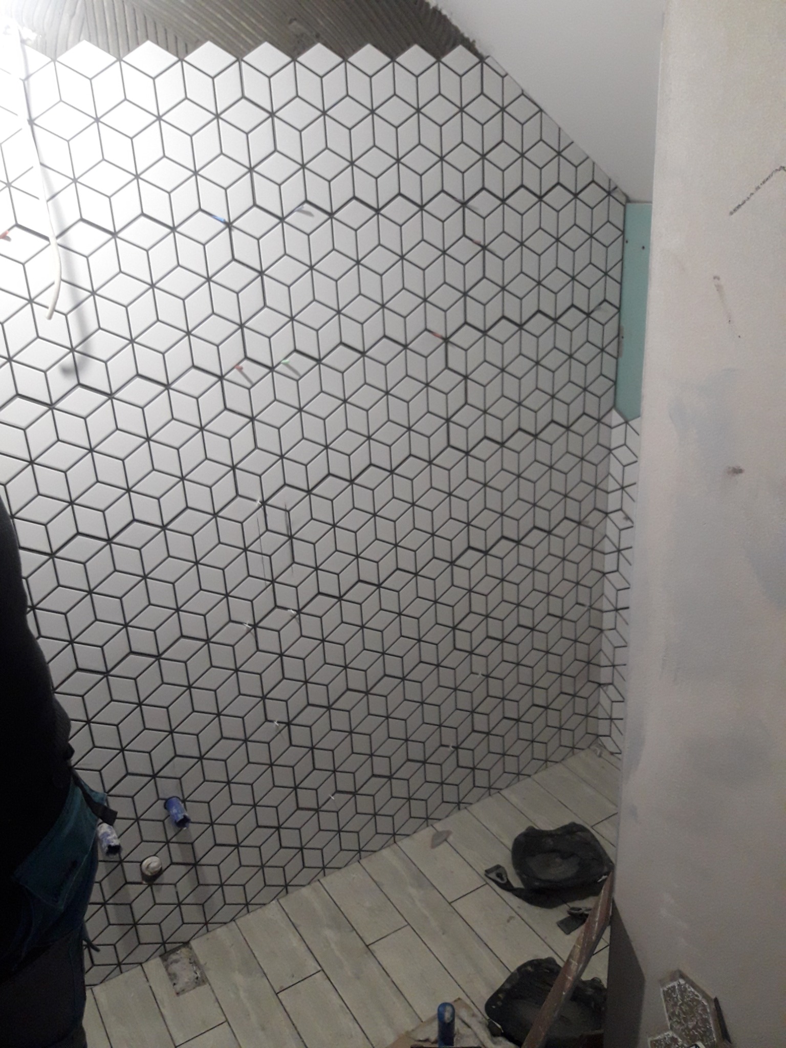 Ściana łazienki w trakcie układania białych płytek o wzorze kostki 3D z ciemnymi fugami, widoczne narzędzia i fragment instalacji hydraulicznej, podłoga wyłożona podłużnymi płytkami imitującymi...