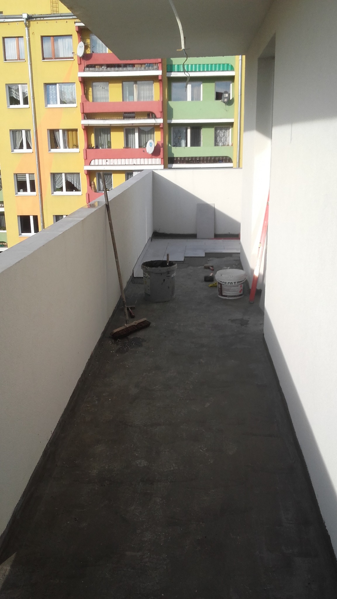 Balkon w trakcie hydroizolacji i układania płytek, widoczne wiadra z materiałami, poziomica i szczotka, w tle kolorowy blok mieszkalny.