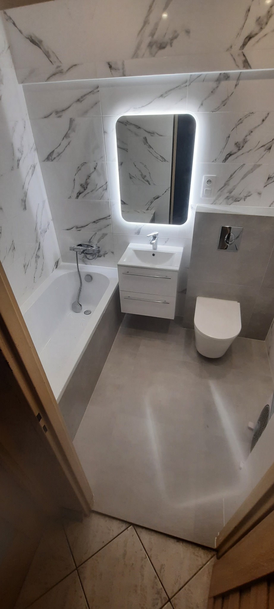 Nowoczesna łazienka z wanną, umywalką z szafką, podświetlanym lustrem i toaletą, wykończona płytkami imitującymi marmur na ścianach i szarymi płytkami na podłodze, widok zza uchylonych drzwi.