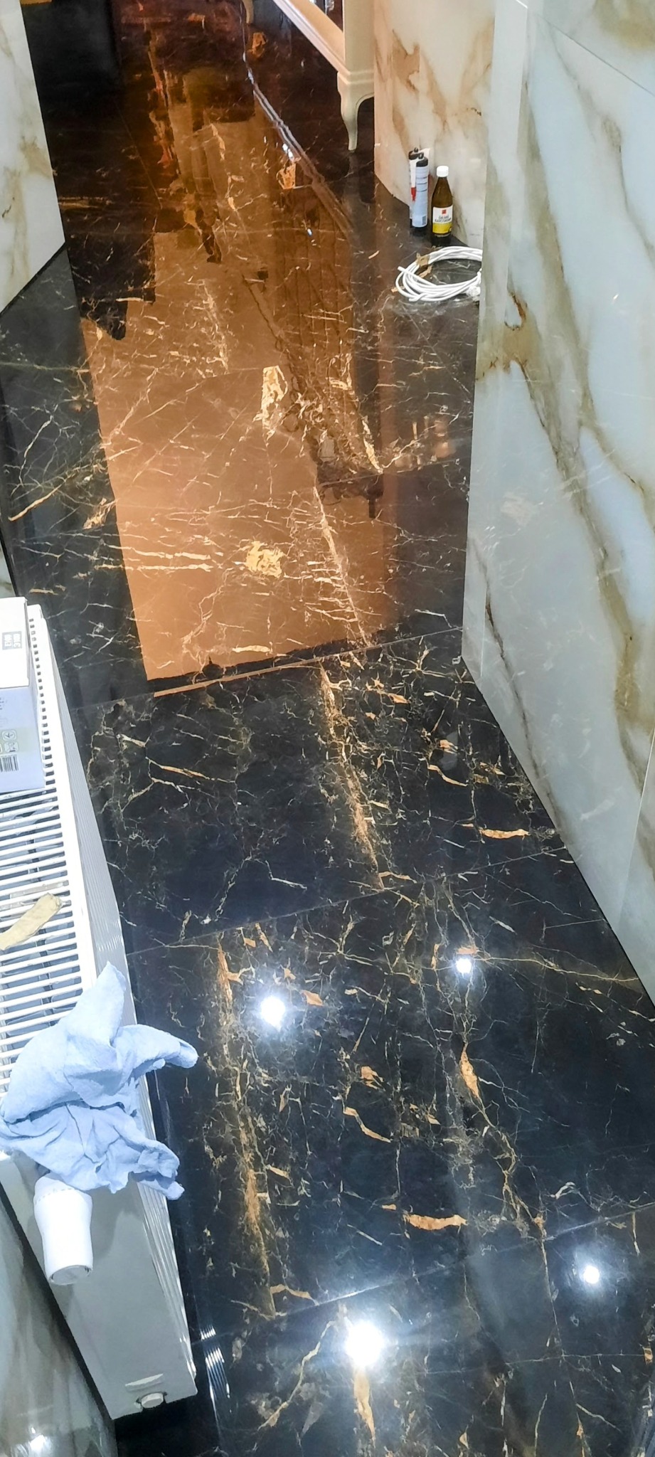 Połyskująca czarna marmurowa podłoga z złotymi żyłami, częściowo wyłożona białymi płytkami imitującymi marmur, widoczne akcesoria glazurnicze i niebieska ścierka na grzejniku.