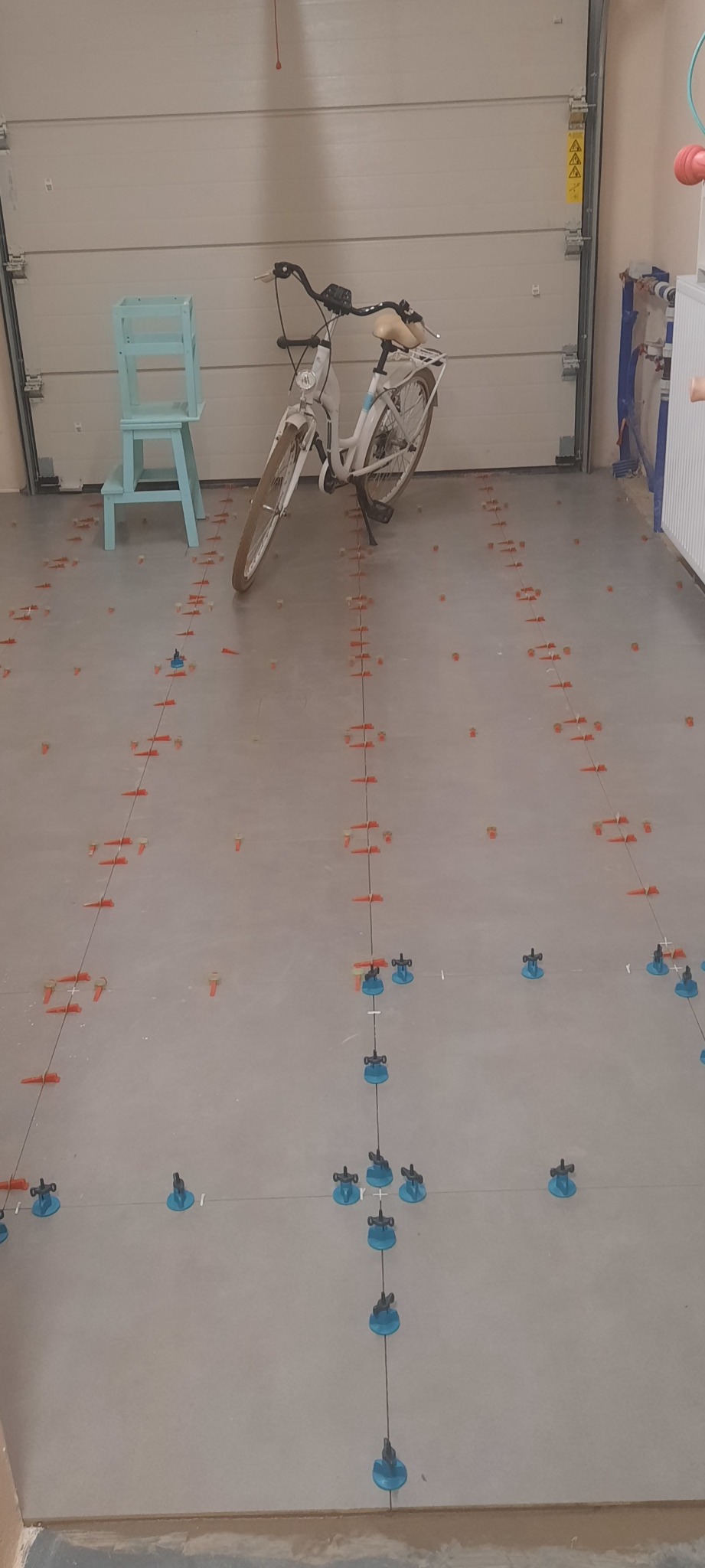 Szare płytki podłogowe w garażu z systemem poziomowania, widoczny rower i krzesło na tle białej bramy garażowej.