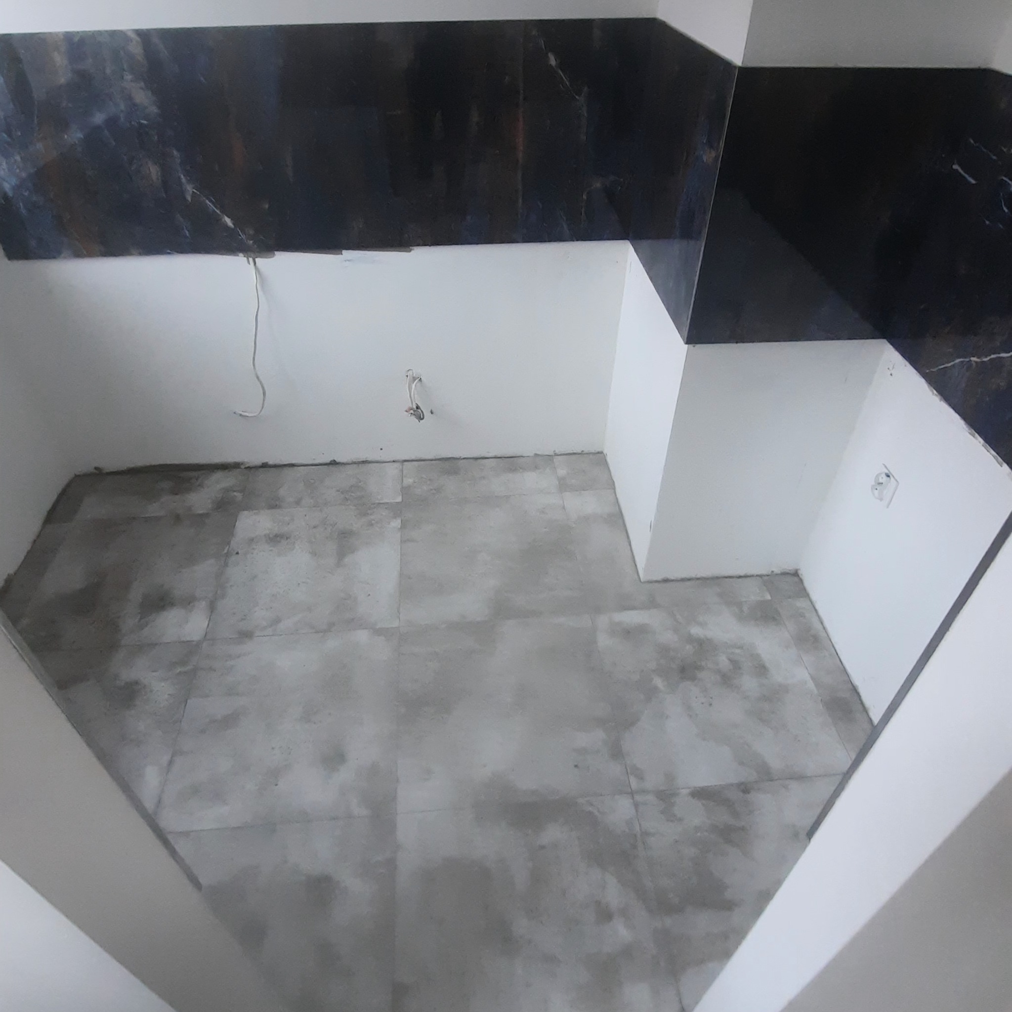 Świeżo położona szara podłoga z płytek w pustym pomieszczeniu, z czarnymi płytkami imitującymi marmur na ścianie, widoczne gniazdko elektryczne i wyprowadzone kable.