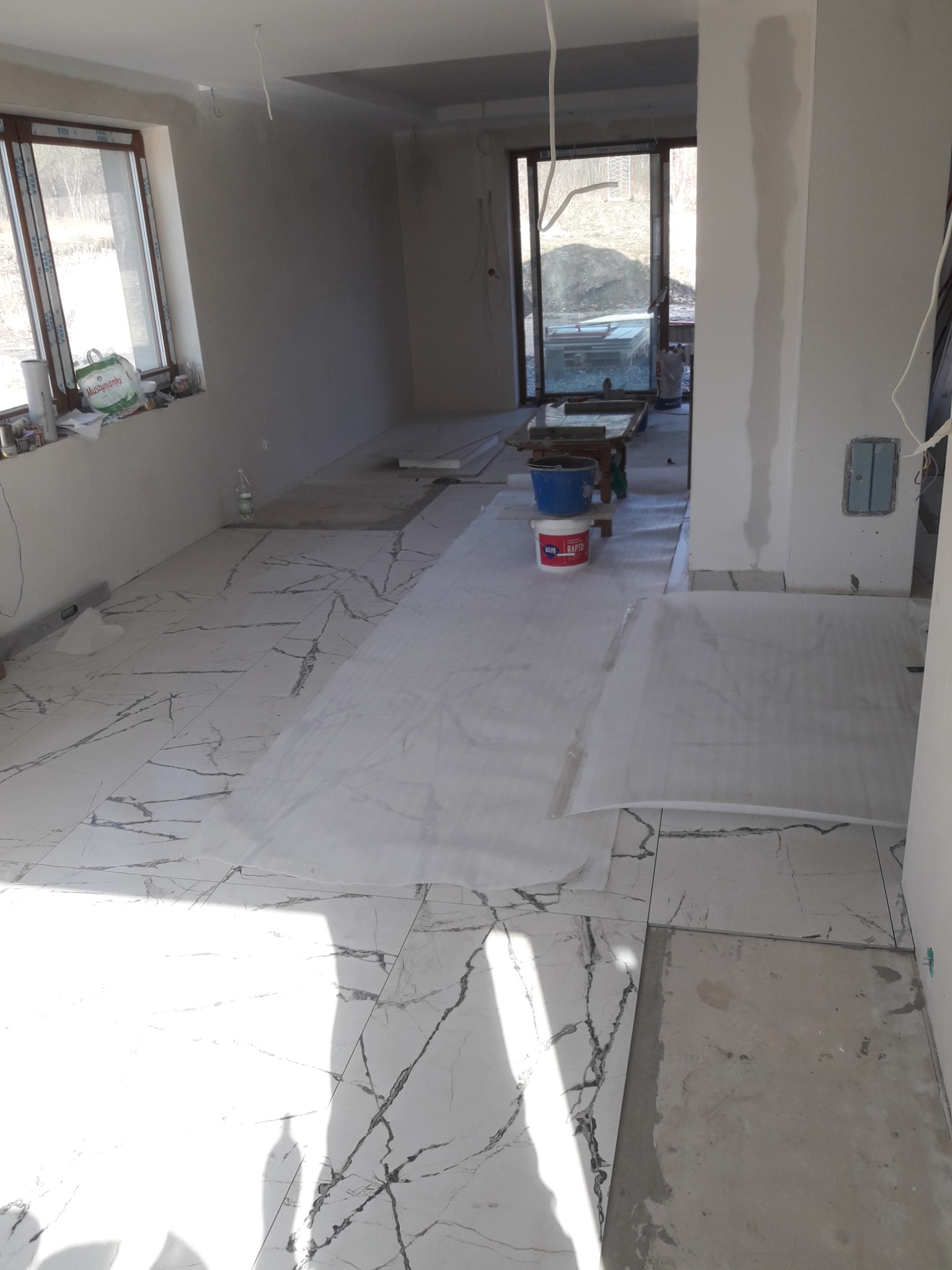 Układanie białych płytek podłogowych imitujących marmur w przestronnym, jasnym pomieszczeniu. Część podłogi zabezpieczona folią ochronną, widoczne wiadro i puszka z klejem do płytek.