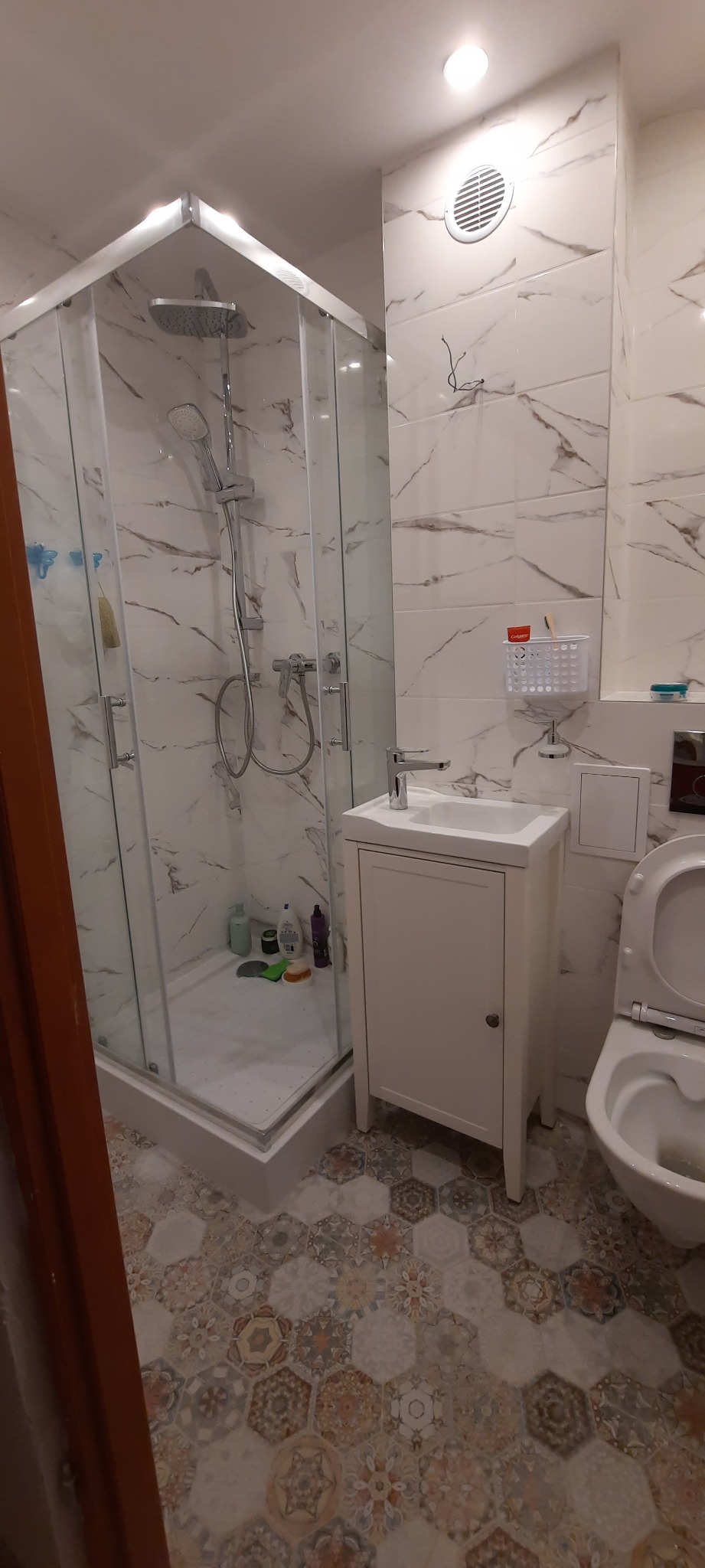 Wykończona łazienka z kabiną prysznicową, umywalką z szafką i toaletą, wyłożona płytkami imitującymi marmur i wzorzystymi heksagonami na podłodze.