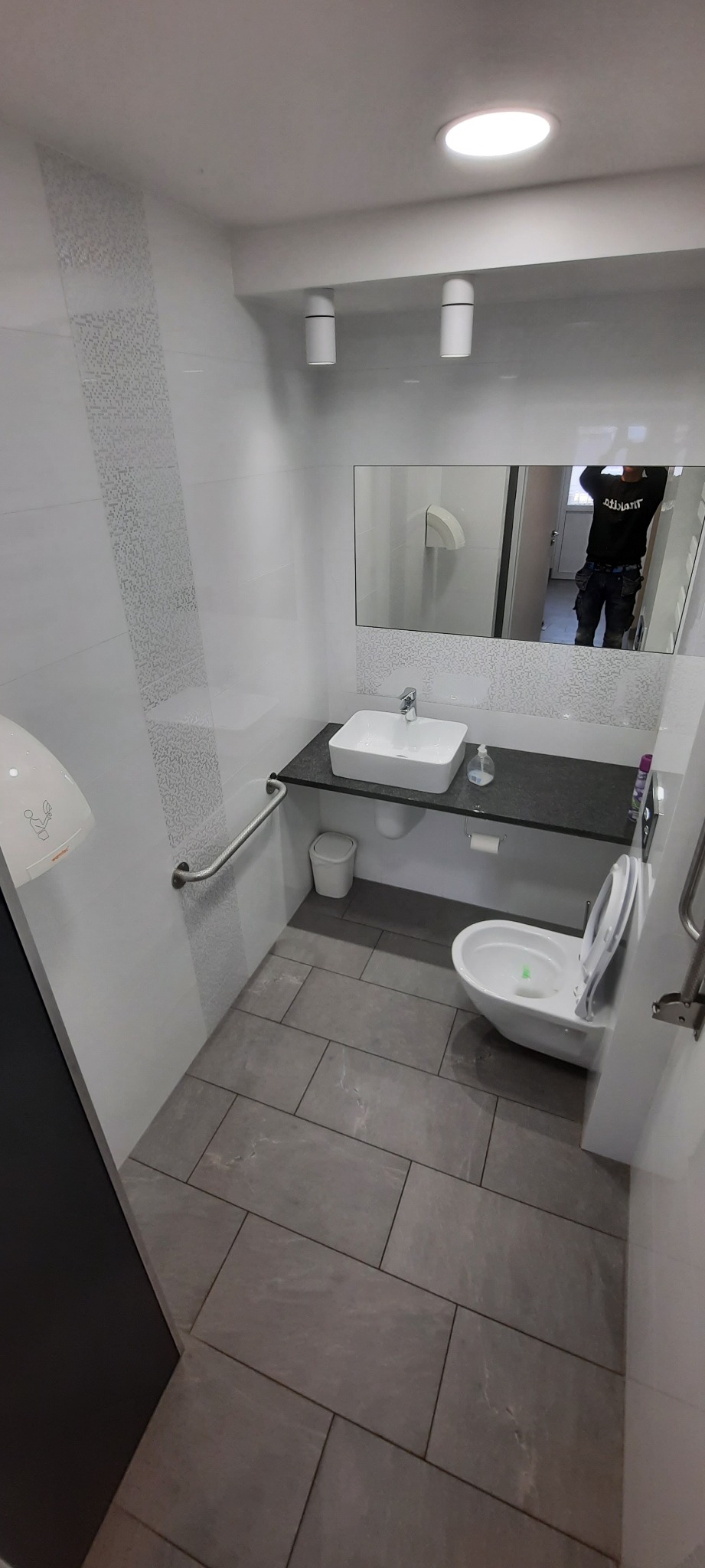 Nowoczesna łazienka z szarymi płytkami podłogowymi, białą ceramiką, armaturą i uchwytem dla niepełnosprawnych. W lustrze odbija się osoba w roboczym ubraniu.