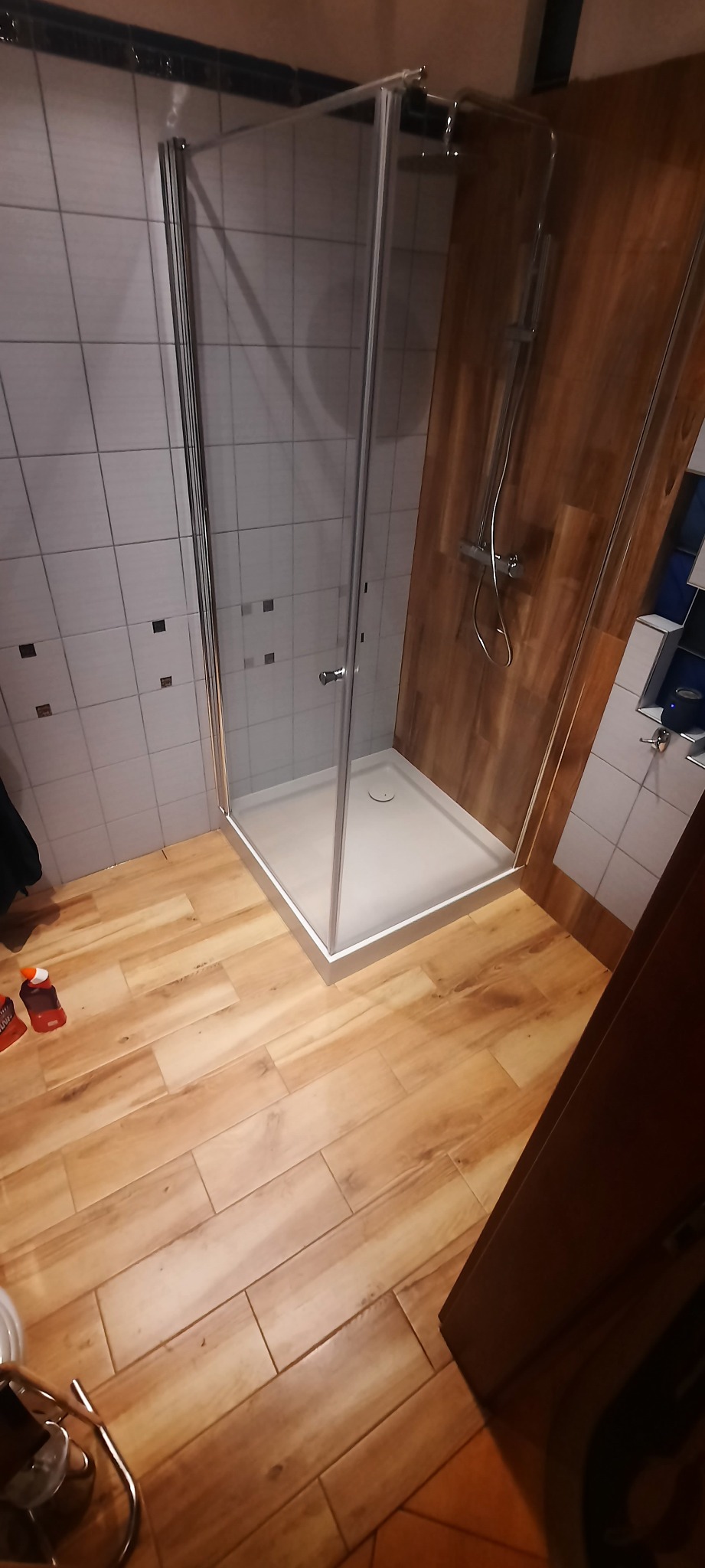 Kabina prysznicowa z chromowanymi profilami na białym brodziku, obok ściana wyłożona płytkami imitującymi drewno, na podłodze płytki drewnopodobne, widoczne środki czyszczące.