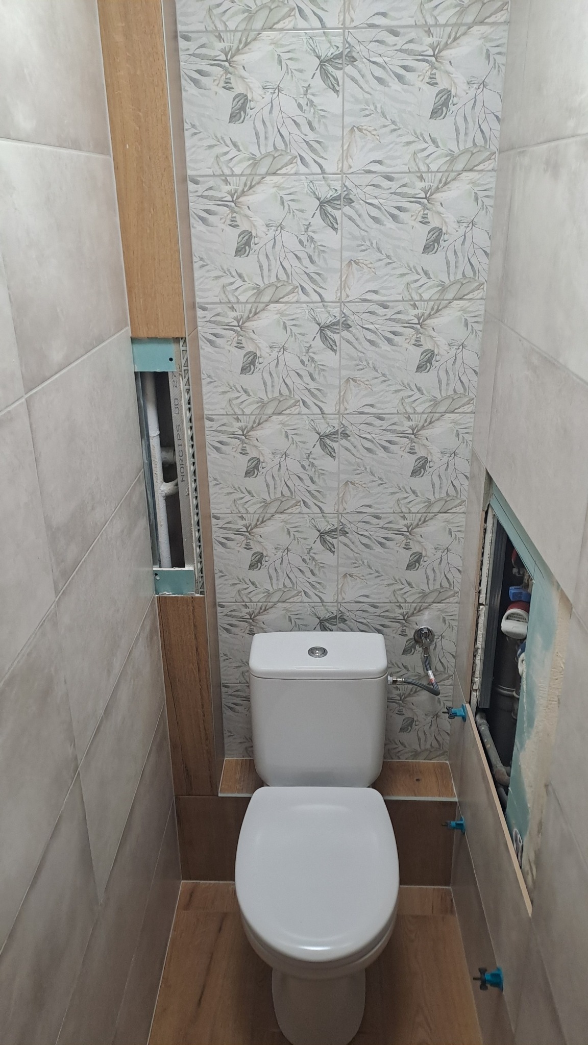 Wnętrze łazienki w trakcie wykańczania z toaletą. Widoczne płytki z motywem roślinnym, fragment niezabudowanej instalacji i niebieskie krzyżyki glazurnicze.