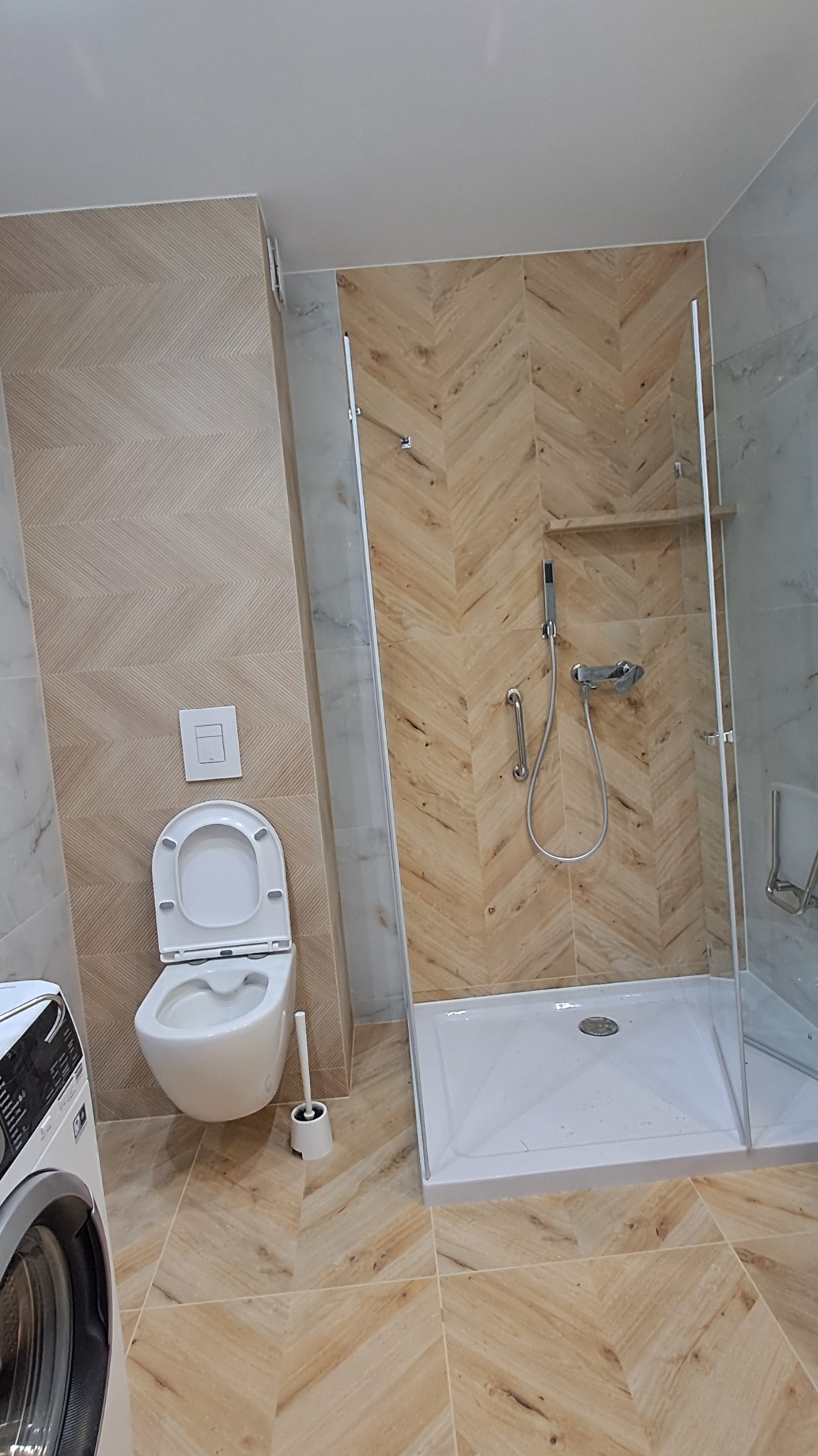 Nowoczesna łazienka z kabiną prysznicową typu walk-in, toaletą podwieszaną i płytkami w jodełkę na ścianach i podłodze. Widoczna pralka i akcesoria łazienkowe.