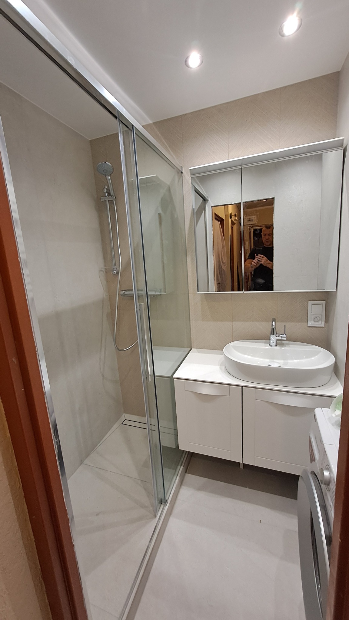 Jasna łazienka z kabiną prysznicową, umywalką nablatową i szafką pod umywalką. W lustrze odbija się osoba robiąca zdjęcie telefonem. Minimalistyczny design.