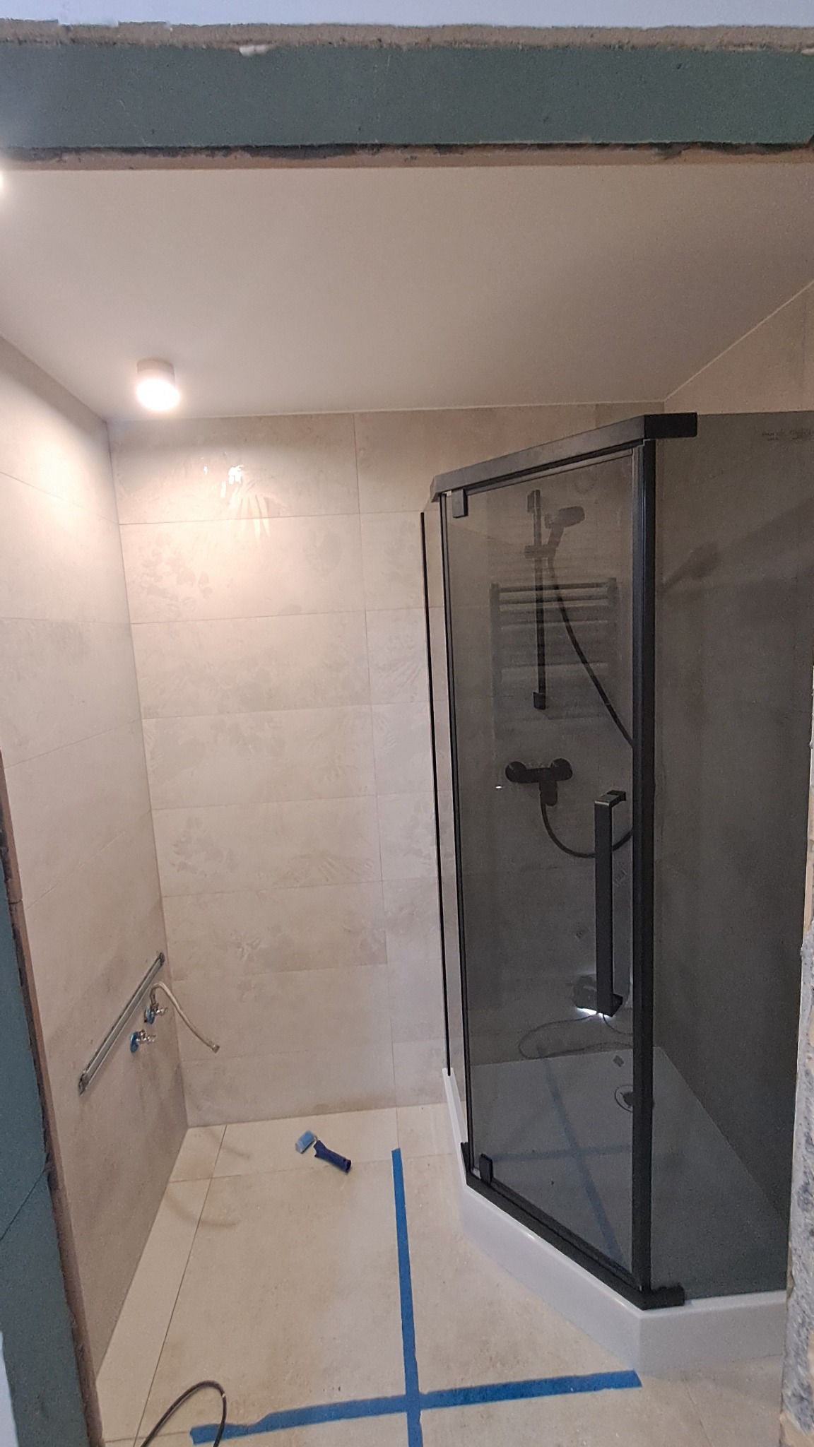 Nowoczesna kabina prysznicowa z czarnymi profilami w jasnej łazience po remoncie, widoczne płytki i narzędzia, taśma malarska na podłodze.