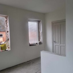 Tomasz Ossowski - Wnętrze po szpachlowaniu ścian na biało. Widoczne okna z ceglanymi wnękami i białe drzwi. Podłoga surowa, przygotowana do dalszych prac wykończeniowych.