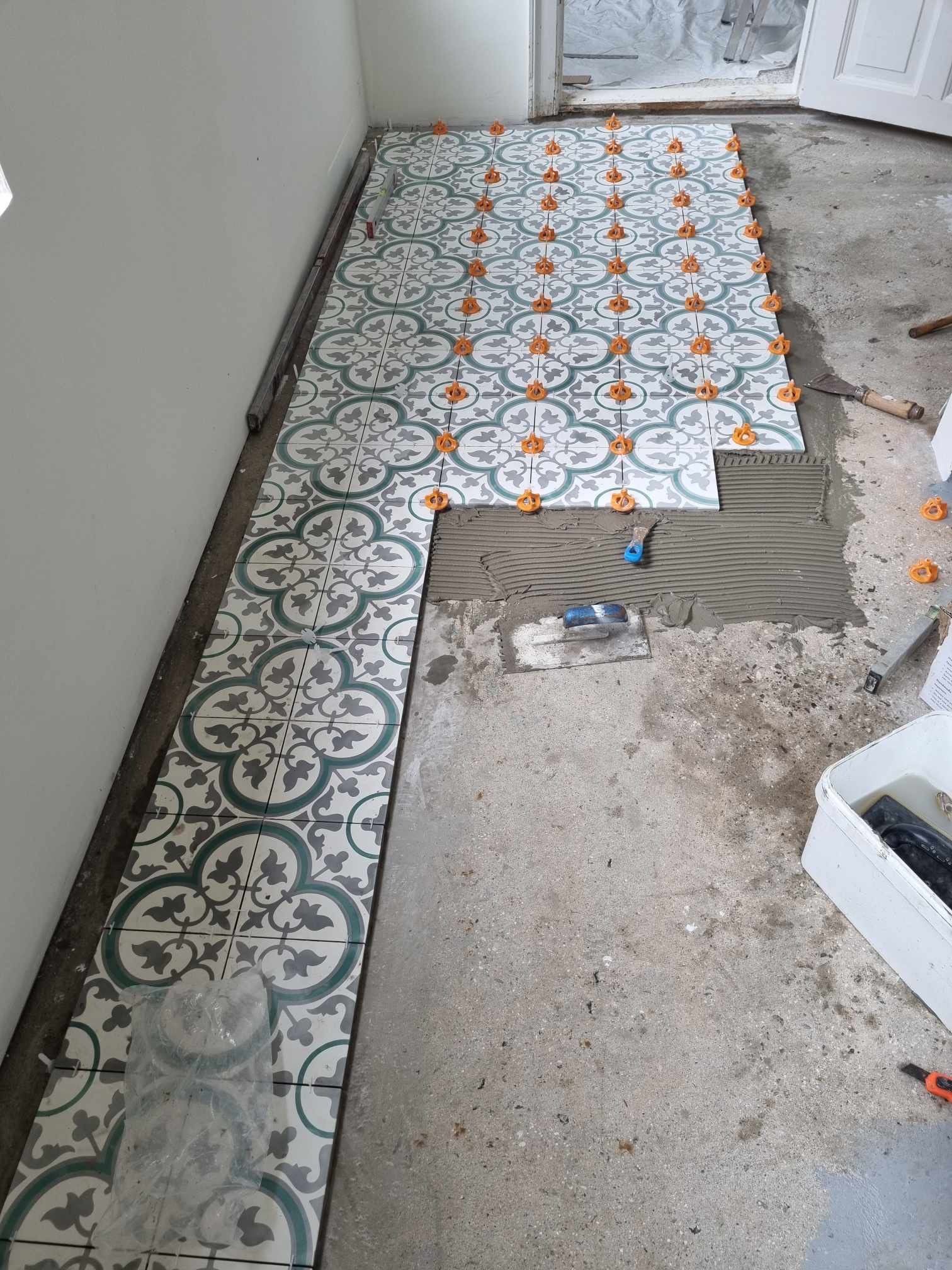 Układanie dekoracyjnych płytek podłogowych z użyciem poziomowania. Widoczna zaprawa, szpachelka i narzędzia. Częściowo ułożona podłoga w pomieszczeniu.