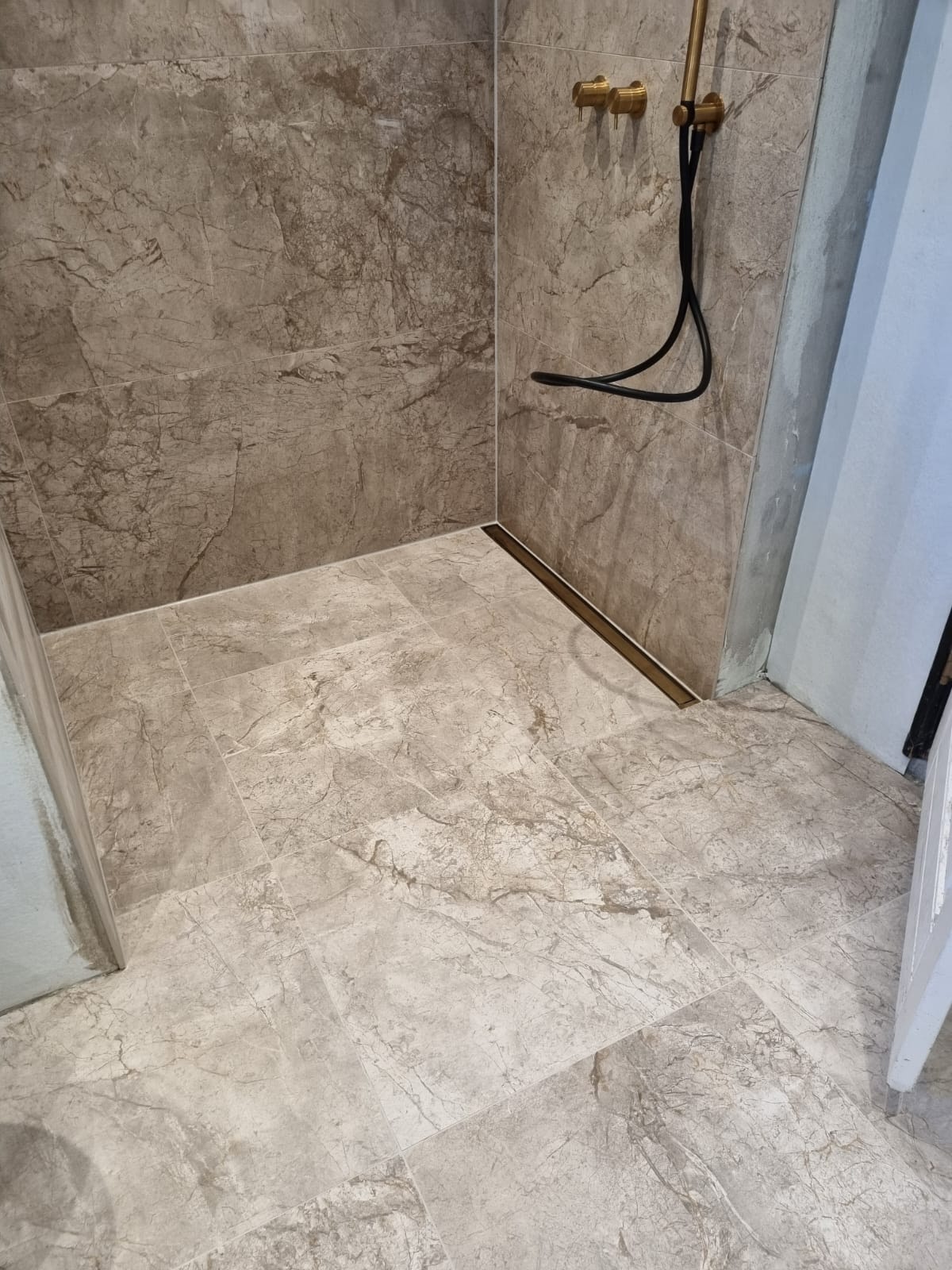 Wykończona kabina prysznicowa z marmurowymi płytkami, złotą armaturą i odpływem liniowym. Widoczne fugi i detale wykończenia.