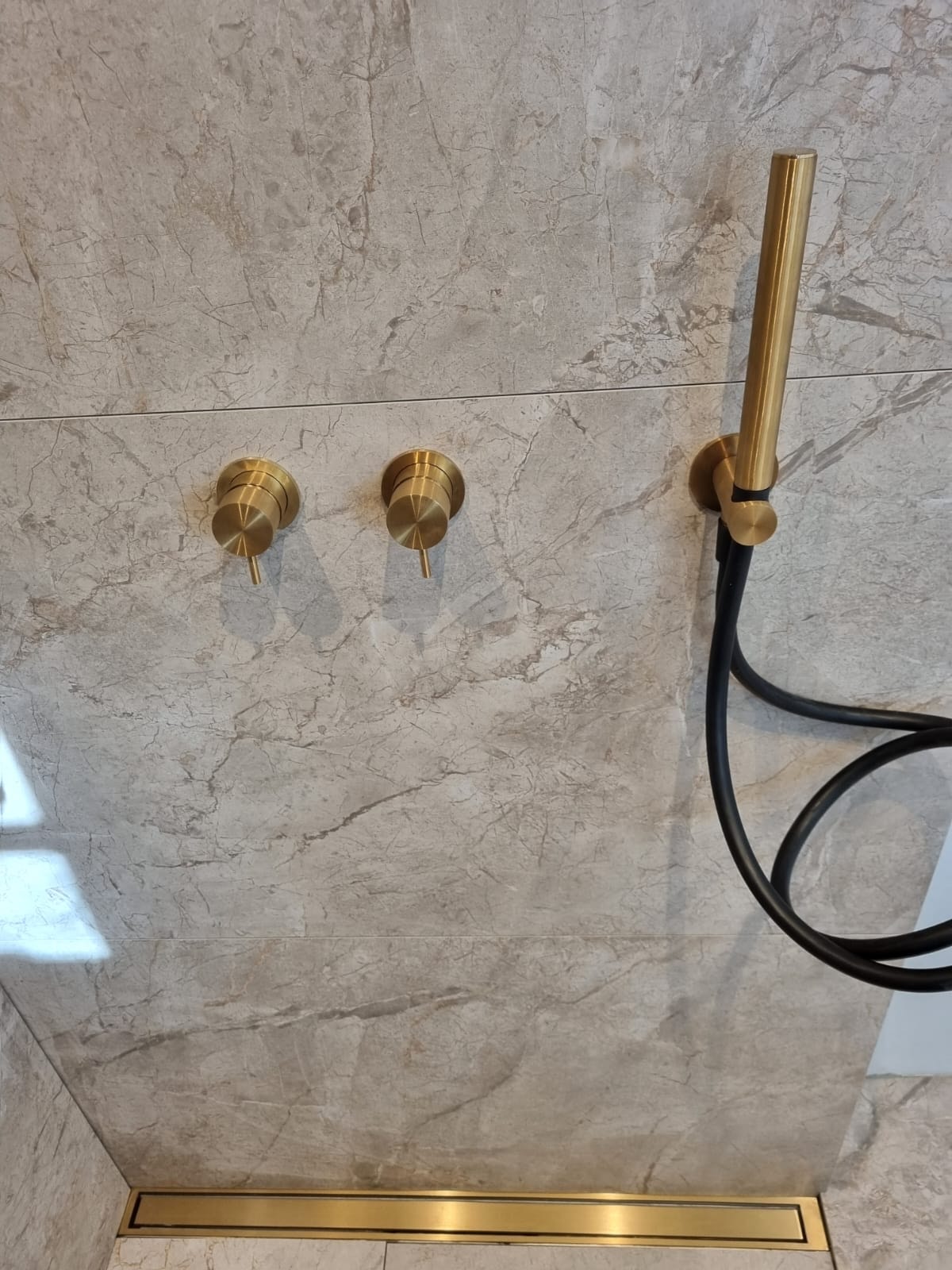 Błyszcząca, złota armatura podtynkowa i odpływ liniowy w łazience wykończonej płytkami imitującymi marmur. Minimalistyczny design, eleganckie detale.