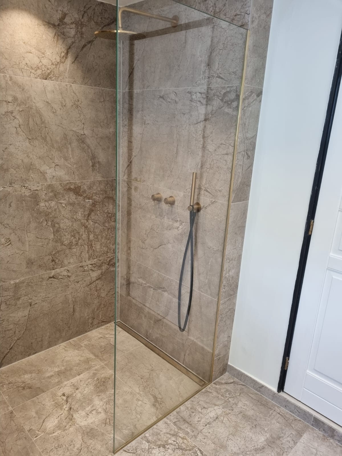 Wykończona kabina prysznicowa walk-in z marmurowymi płytkami w odcieniach beżu i brązu. Złote wykończenia armatury i ramy kabiny, szklana tafla bez brodzika.