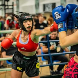 Darka1 Fatburner - Kobieta w czerwonym topie i ochraniaczu na głowę zadaje cios w ringu podczas walki, widoczni widzowie w tle.