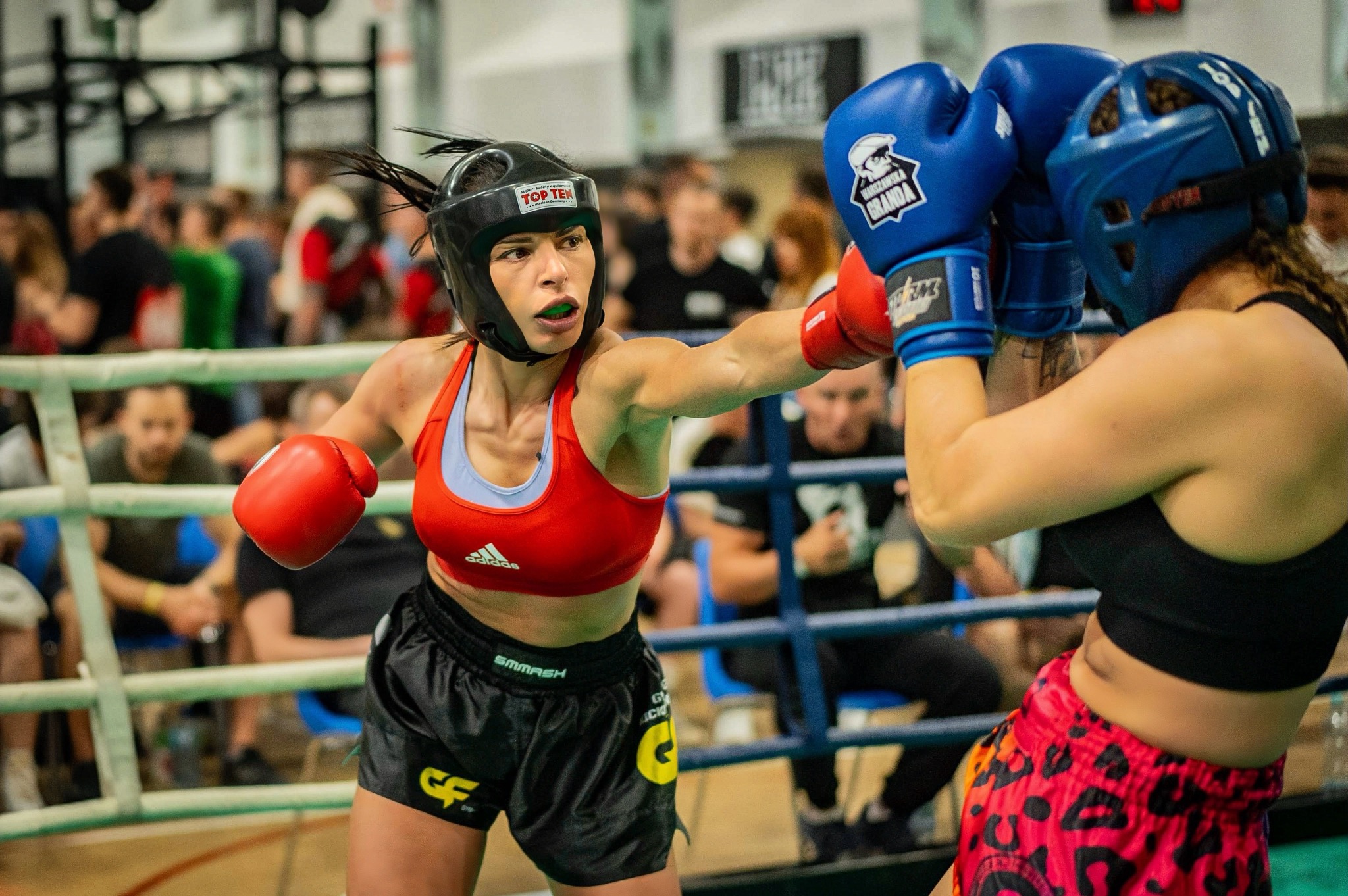 Kobieta w czerwonym topie i ochraniaczu na głowę zadaje cios w ringu podczas walki, widoczni widzowie w tle.