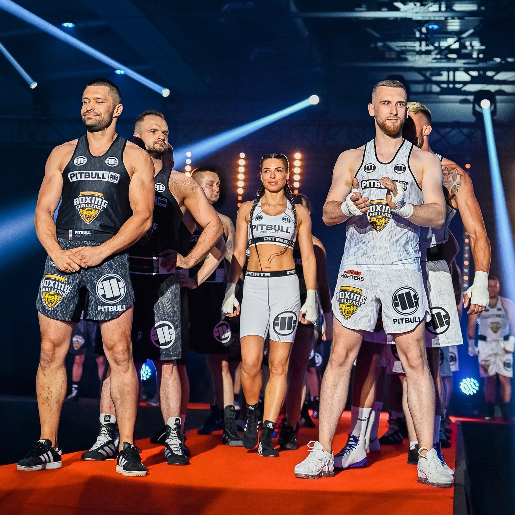 Grupa zawodników sportów walki w strojach sportowych z logo marki Pitbull na scenie oświetlonej reflektorami podczas wydarzenia sportowego.