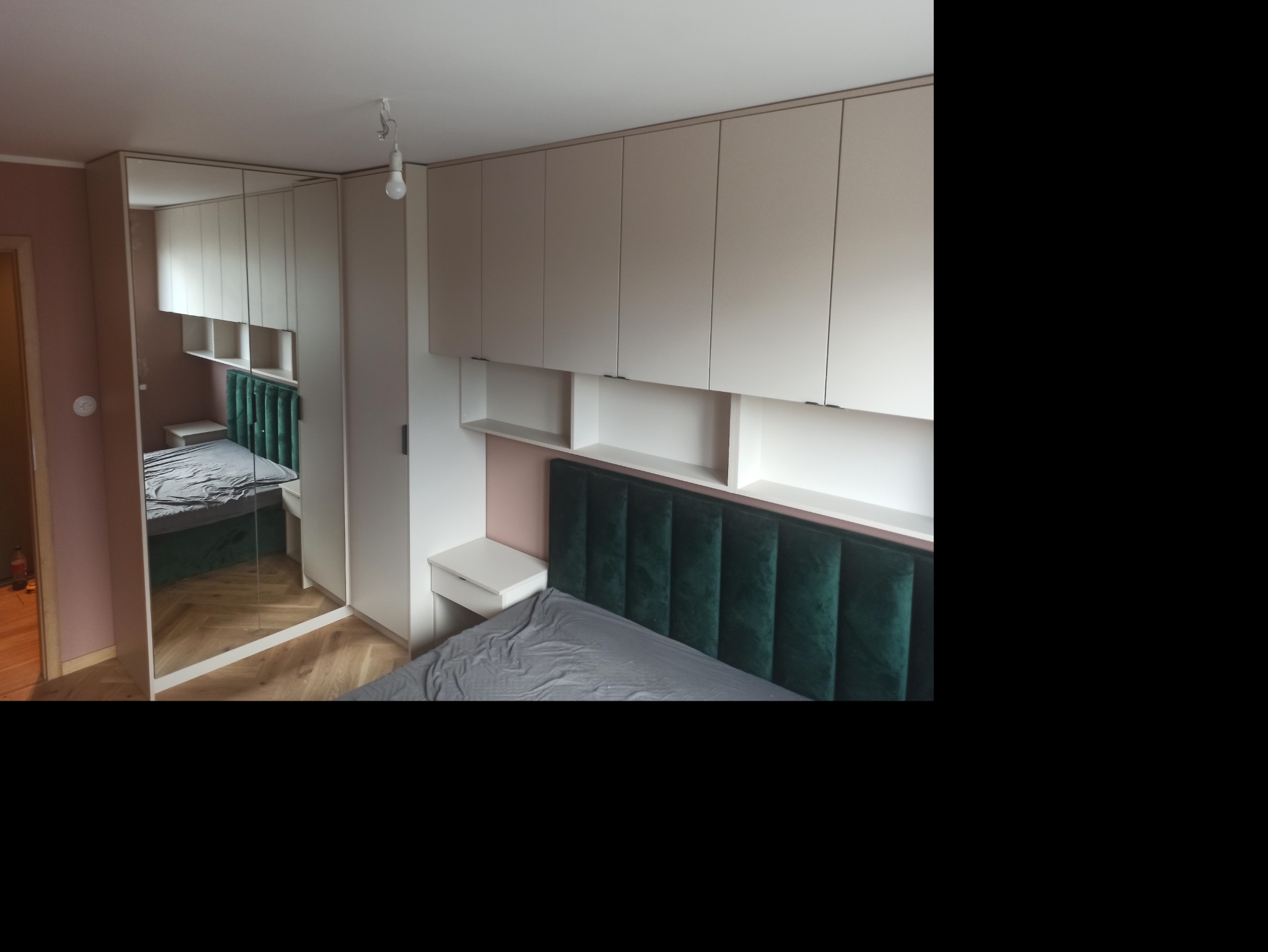 Szafa wnękowa z lustrzanymi drzwiami w sypialni, zintegrowana z szafkami nad łóżkiem z zielonym zagłówkiem, widoczne odbicie parkietu.