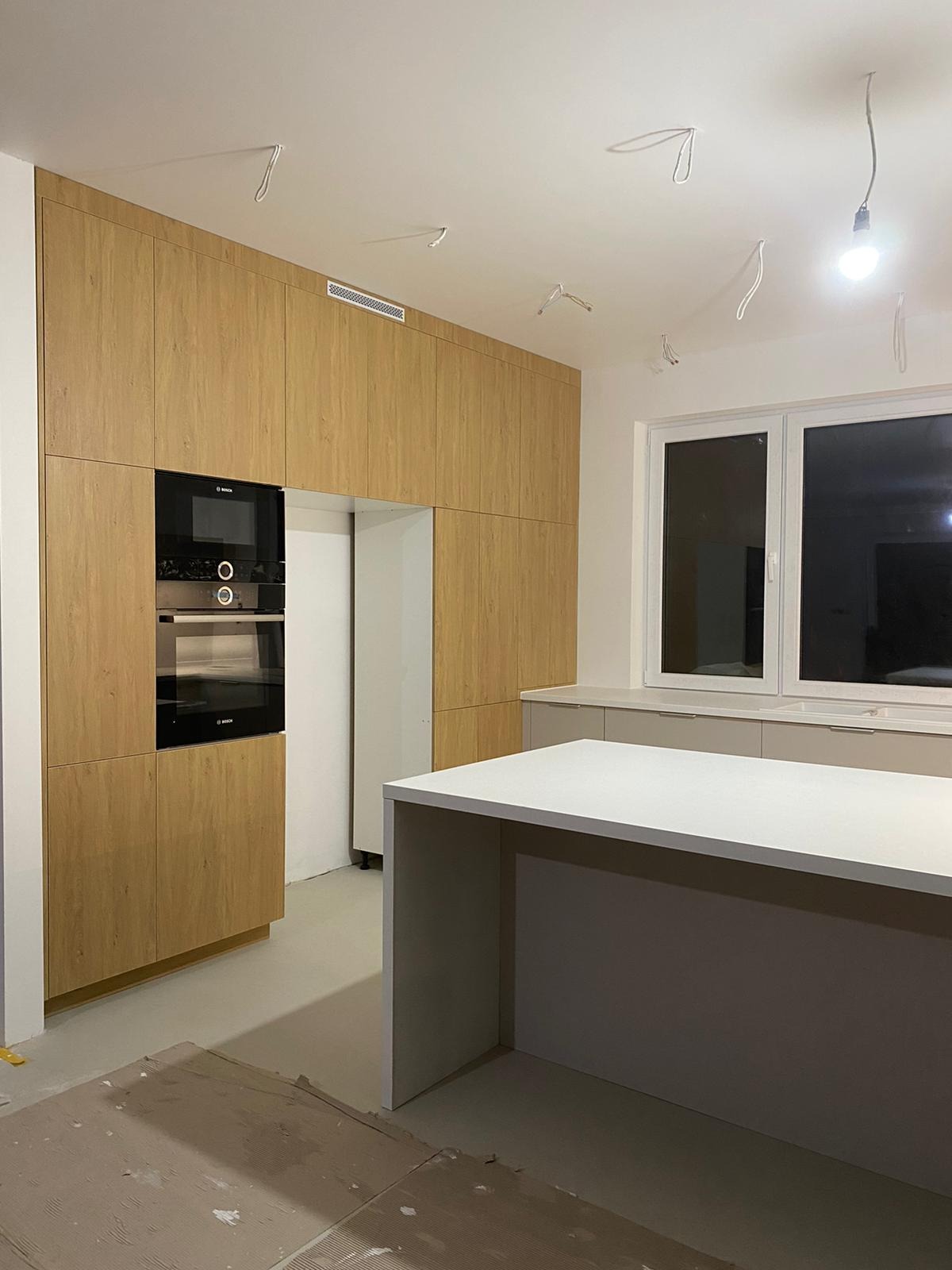 Nowoczesna kuchnia w trakcie montażu: zabudowa z drewnianymi frontami, wbudowany piekarnik i płyta, wyspa kuchenna z białym blatem, surowe wykończenie podłogi i wiszące kable.