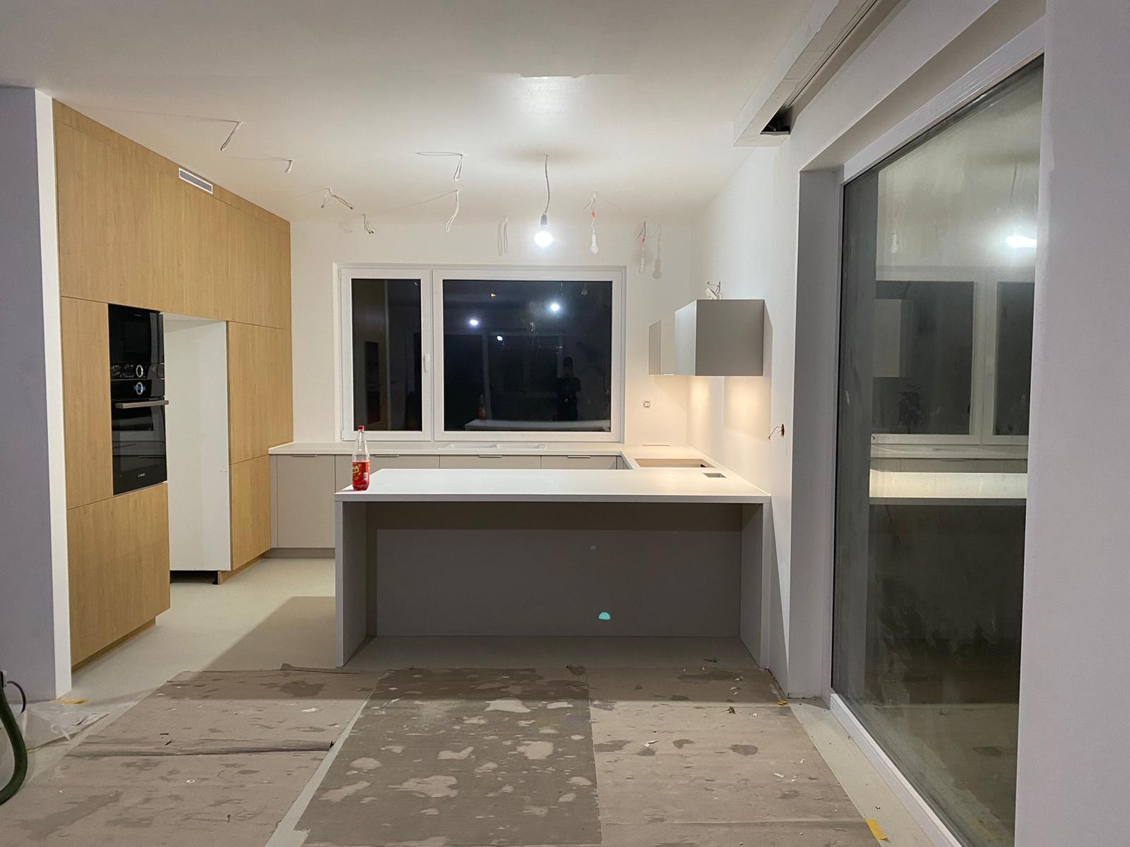 Wnętrze kuchni w trakcie montażu, widoczne drewniane szafki, biały blat roboczy, okno z widokiem na zewnątrz, podłoga zabezpieczona kartonem, wiszące kable od oświetlenia.