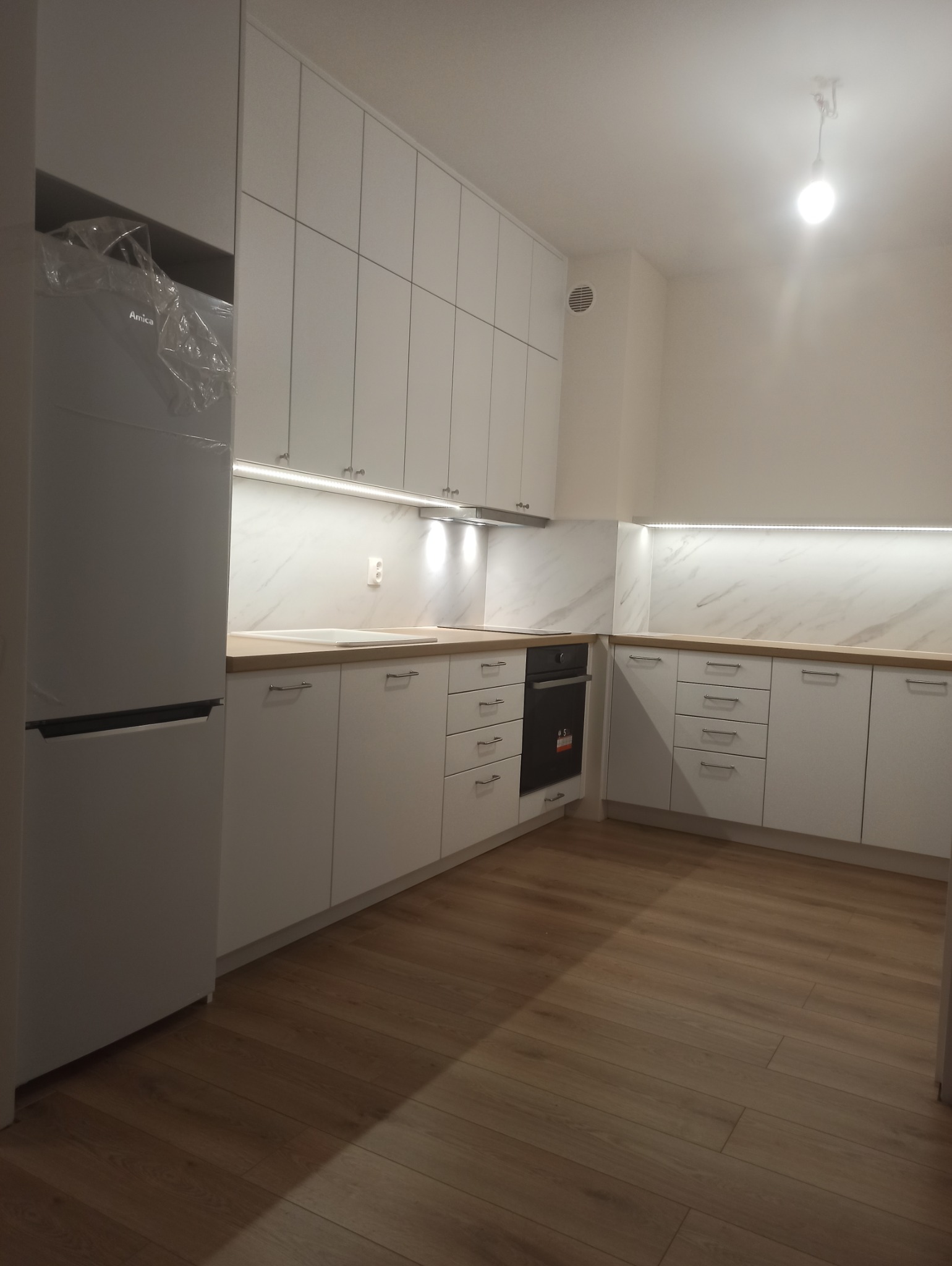 Jasna, nowoczesna kuchnia z białymi szafkami, drewnianym blatem, marmurowym backsplashiem i nową lodówką Amica w folii ochronnej.