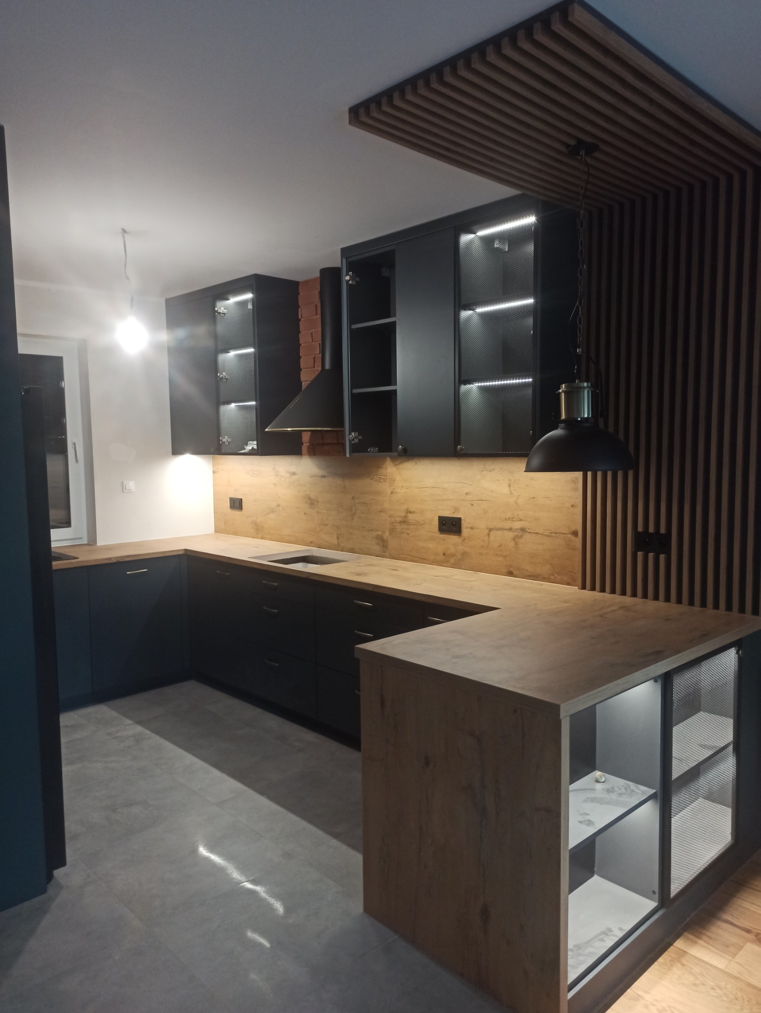 Nowoczesna kuchnia z czarnymi szafkami z oświetleniem LED, drewnianym blatem i panelem ściennym imitującym drewno, wykończona ceglanym kominem i dekoracyjnymi lamelami na suficie i ścianie.