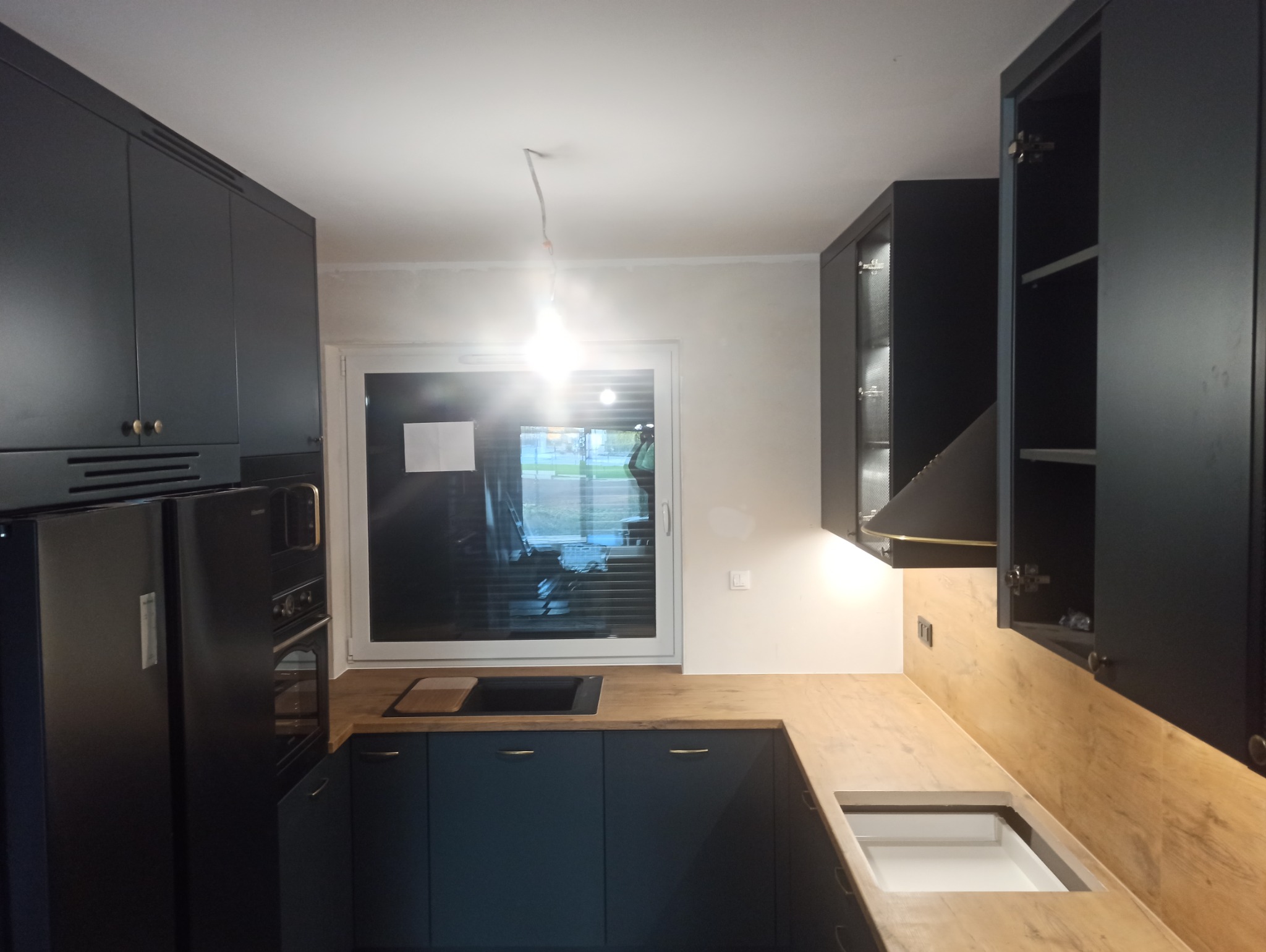 Nowoczesna kuchnia z ciemnymi, matowymi szafkami, drewnianym blatem i czarnym sprzętem AGD, widoczne niedokończone prace wykończeniowe przy suficie i oświetleniu.