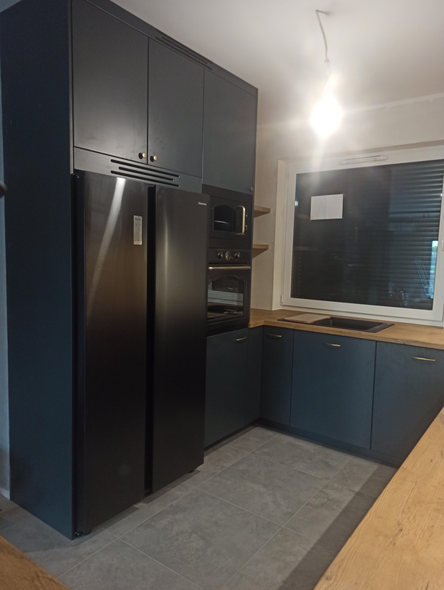 Nowoczesna kuchnia w odcieniach grafitu i drewna: zabudowa kuchenna z szafkami górnymi, lodówka side-by-side, piekarnik i płyta grzewcza w zabudowie, drewniany blat, szare płytki podłogowe.