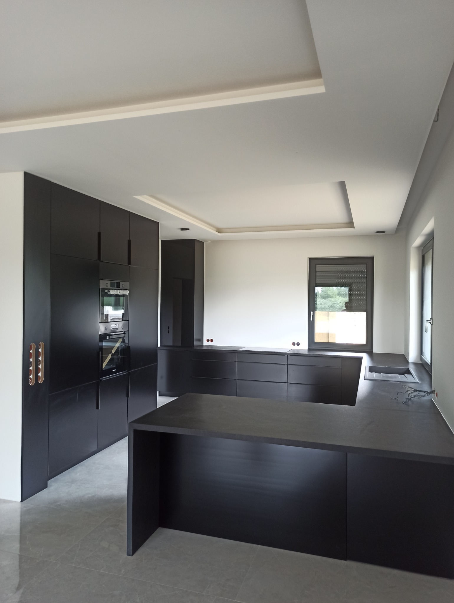 Nowoczesna kuchnia w kolorze antracytowym z wyspą kuchenną i zabudową pod sufit, minimalistyczne uchwyty, szare płytki podłogowe.