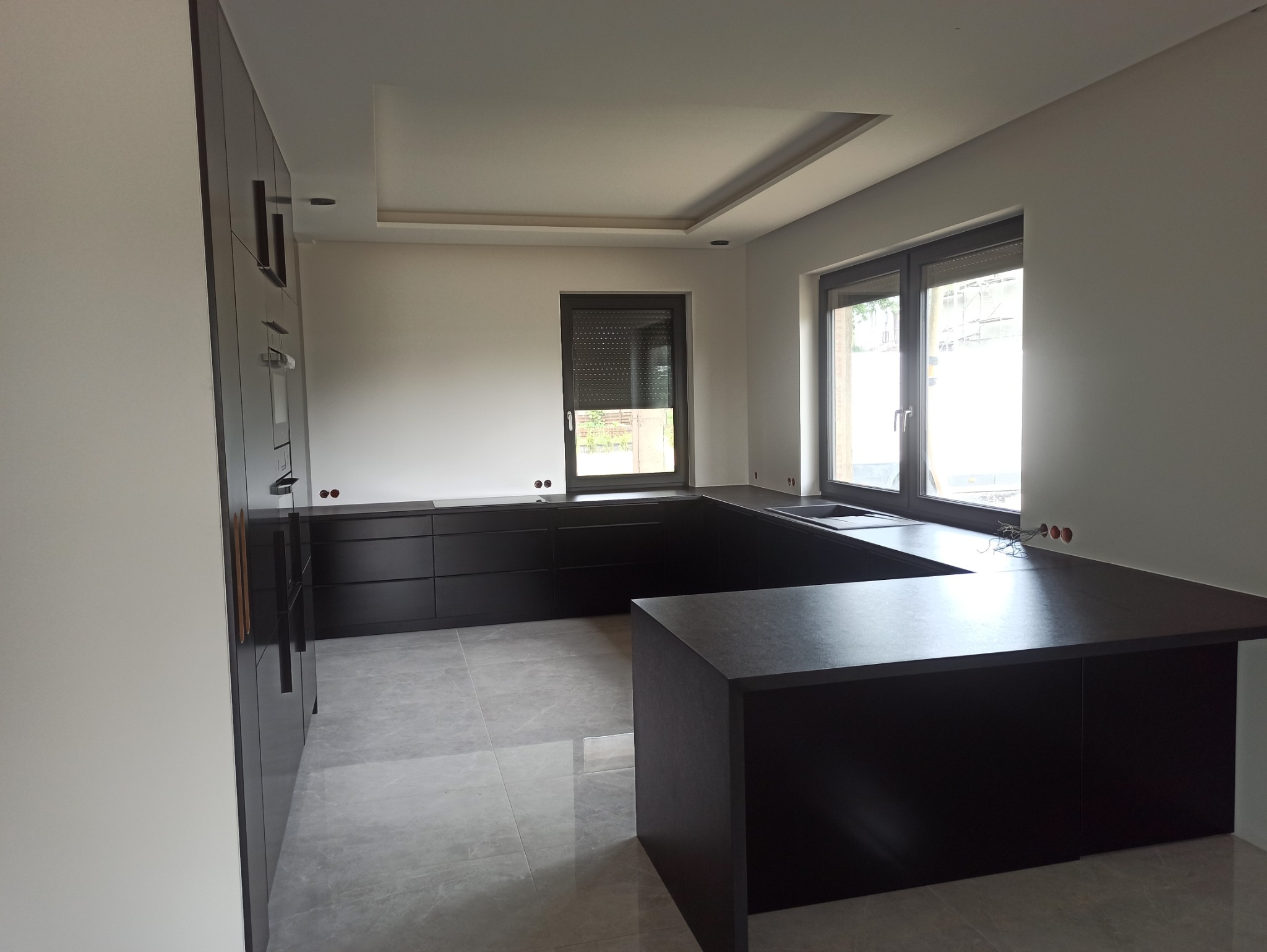 Nowoczesna kuchnia w ciemnej kolorystyce z szarymi płytkami podłogowymi i widocznymi instalacjami elektrycznymi na ścianie.