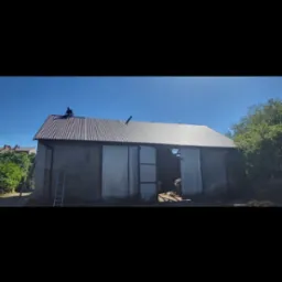 Pracownik na dachu krytym blachą trapezową, drabina oparta o ścianę budynku z szarego pustaka, brama garażowa z blachy falistej, widok na wieś w tle.